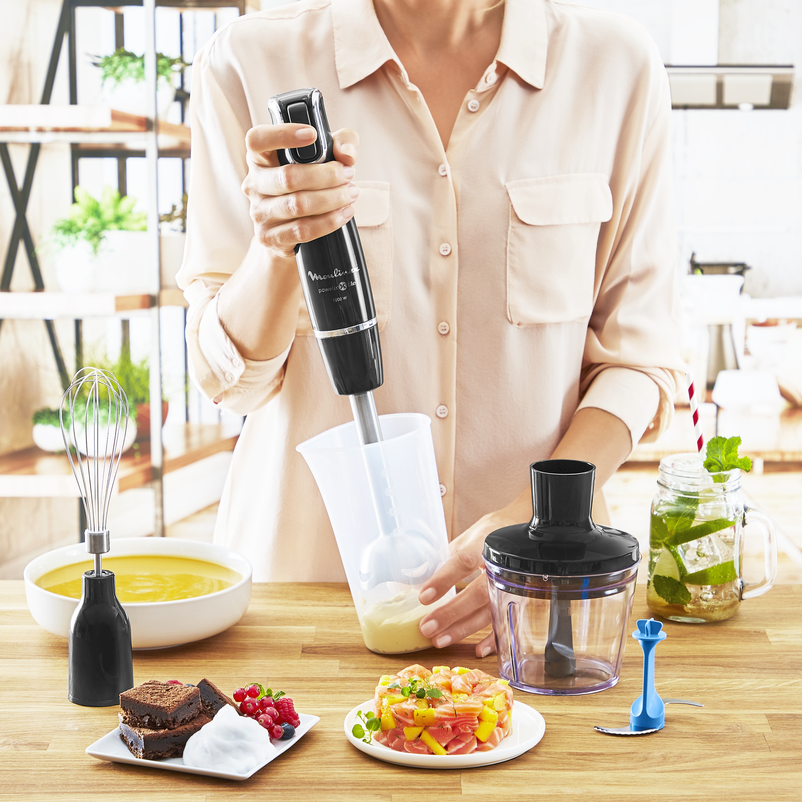 A person using a black hand blender to blend ingredients in a clear container.