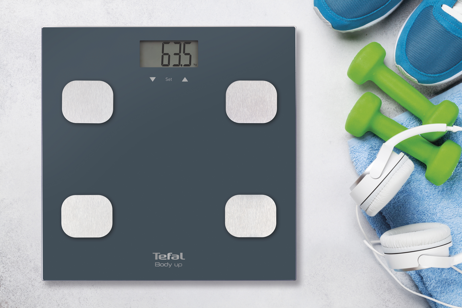 Une balance numérique affiche 63,5 kg. À côté, haltères, écouteurs et baskets sur une serviette bleue.