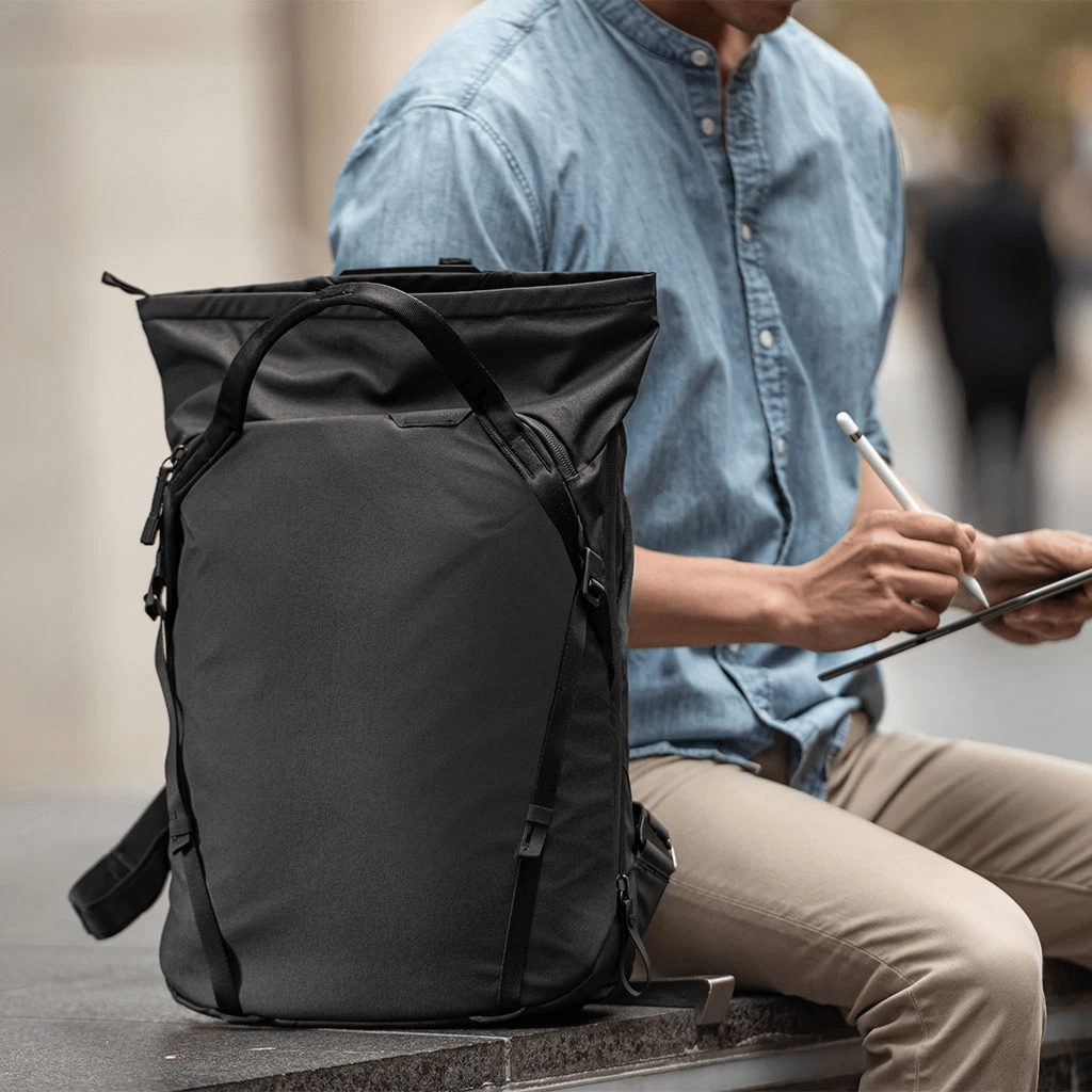 Uno zaino nero su una superficie di cemento. Un uomo, con un tablet, è seduto vicino.