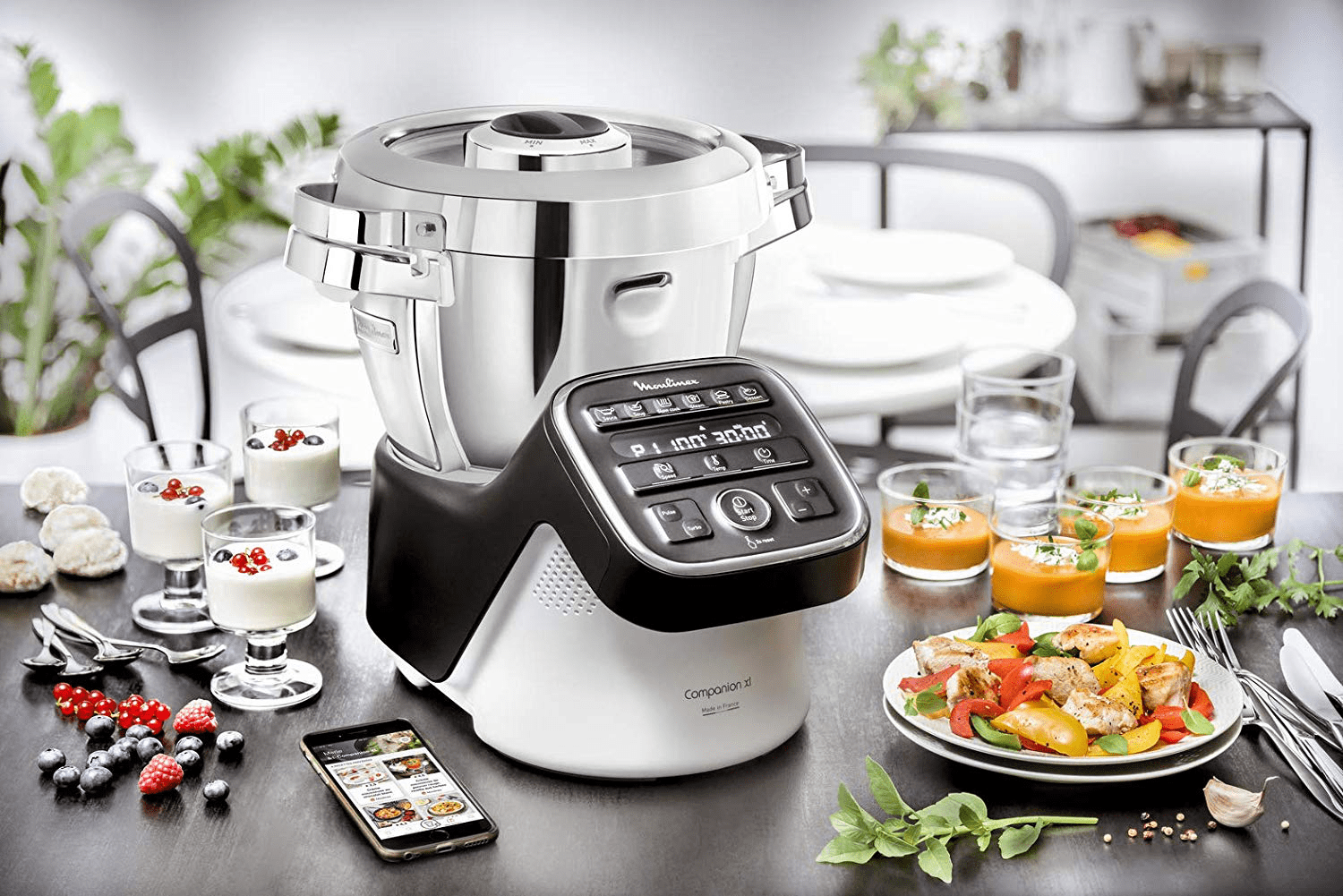 Scène de préparation culinaire : un appareil de cuisine avec de la nourriture et des boissons sur une table et un smartphone.