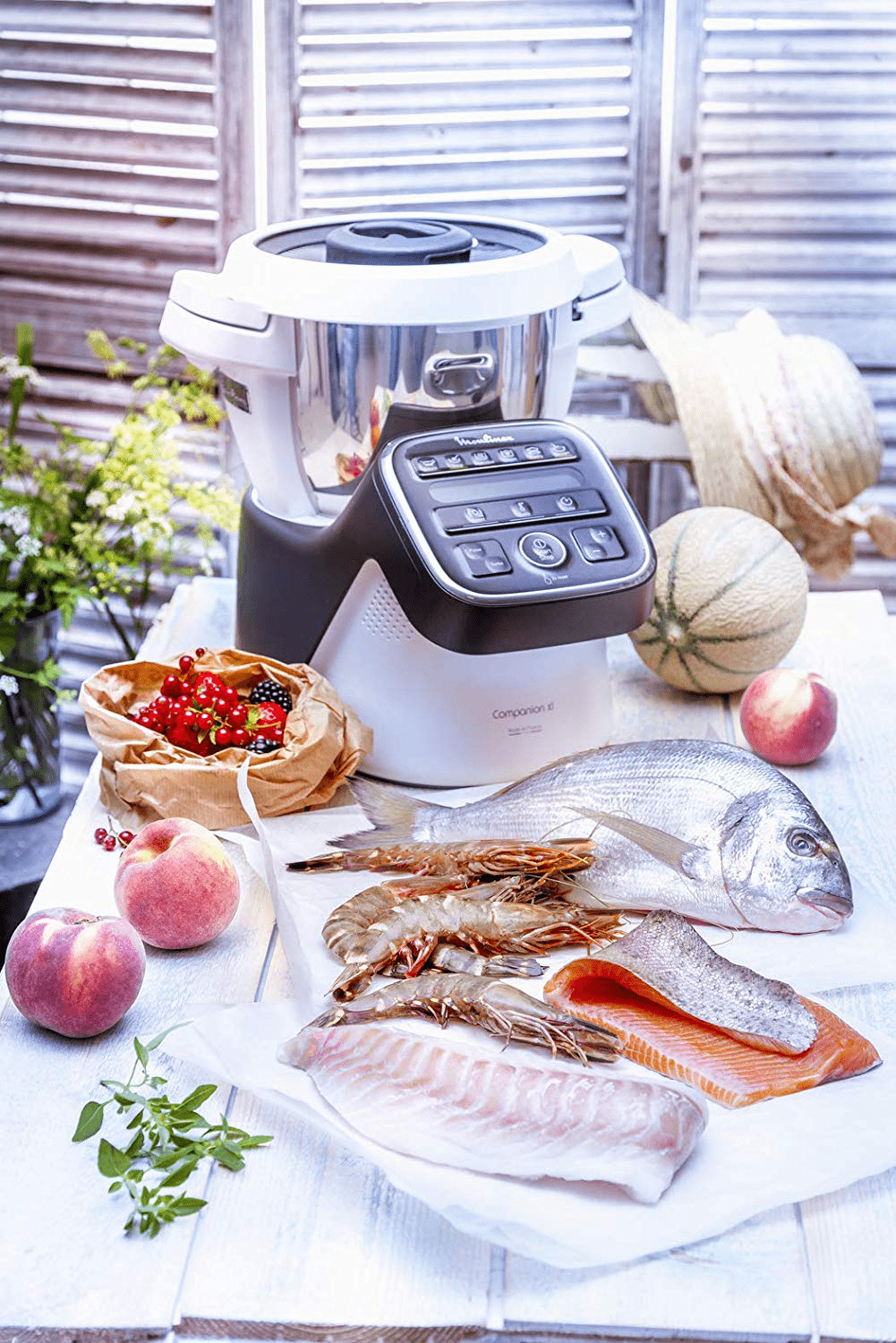 Une scène de préparation culinaire avec un robot culinaire, du poisson cru, des crevettes, des pêches et des baies sur une table.