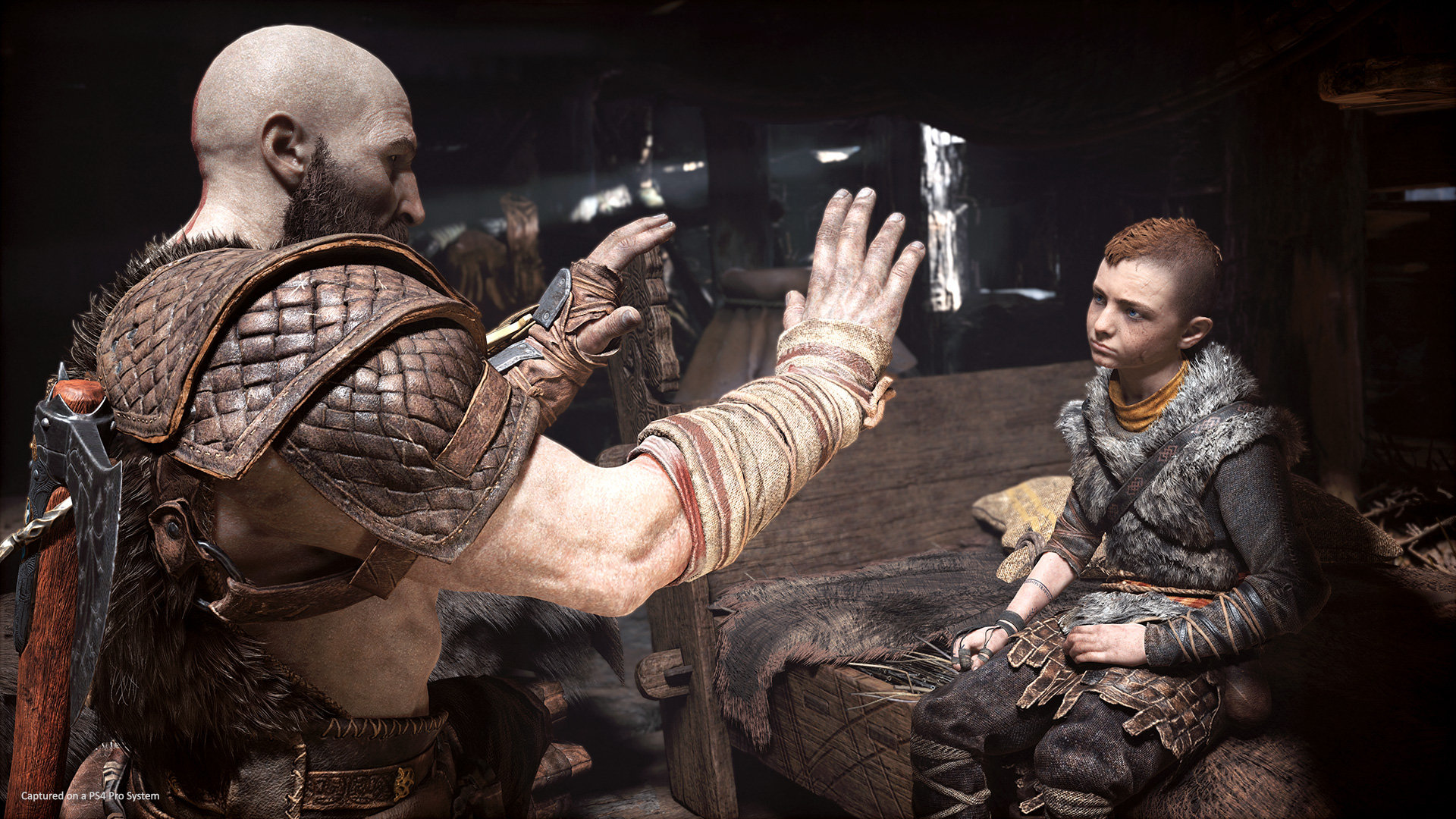Kratos y Atreus en un interior con poca luz, Kratos está gesticulando hacia Atreus.