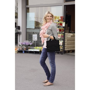 Eine blonde Frau im grauen Cardigan und rosa Schal steht mit einer schwarzen Handtasche vor einem Geschäft.