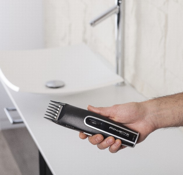 La mano de una persona sosteniendo una cortadora de cabello Rowenta negra y plateada cerca de un lavabo blanco.