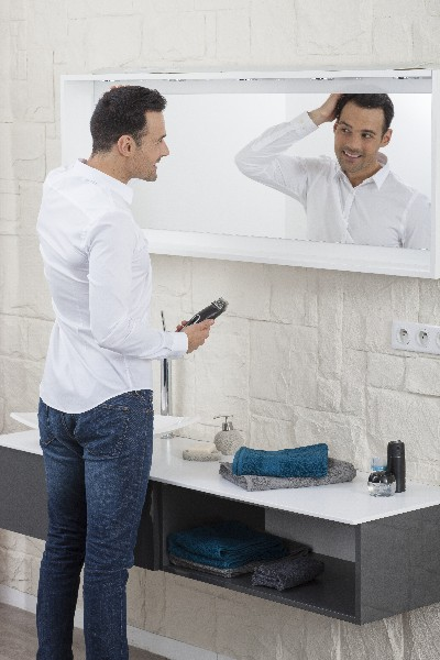 Hombre mirándose en el espejo, sosteniendo una recortadora en el baño. Pantalón vaquero y camisa blanca.