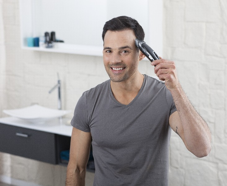 Un hombre sonríe mientras sostiene una cortadora de pelo. Está en un baño, y el fondo es un espejo.