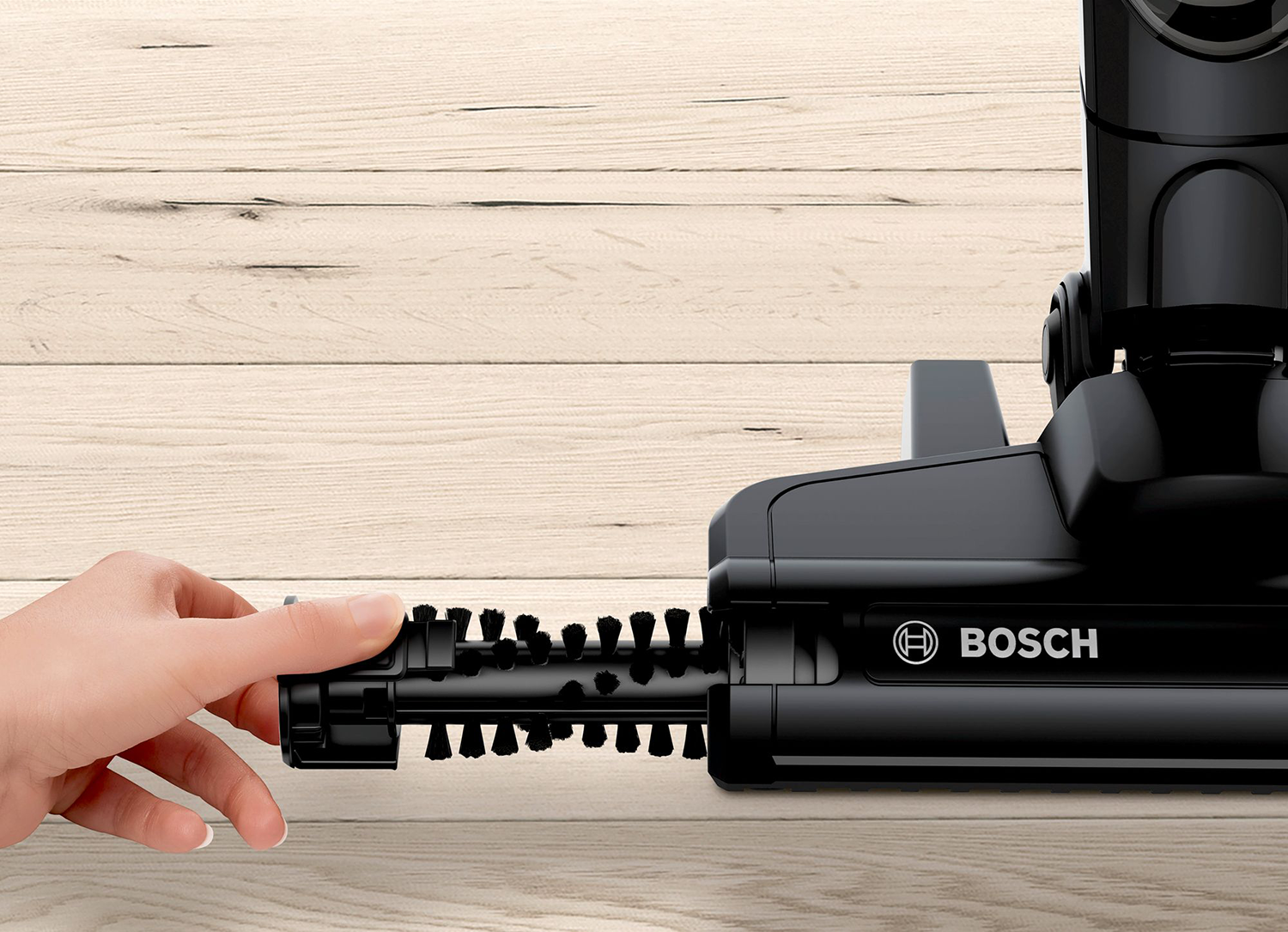 A hand holding a brush on a black vacuum cleaner brush head, on a wooden surface.