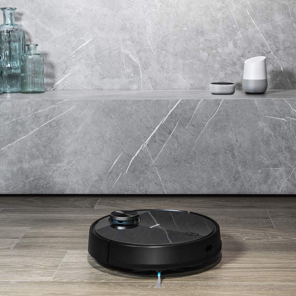 Aspiradora robot negra en un piso de madera. Una pared de mármol gris con decoración está detrás.
