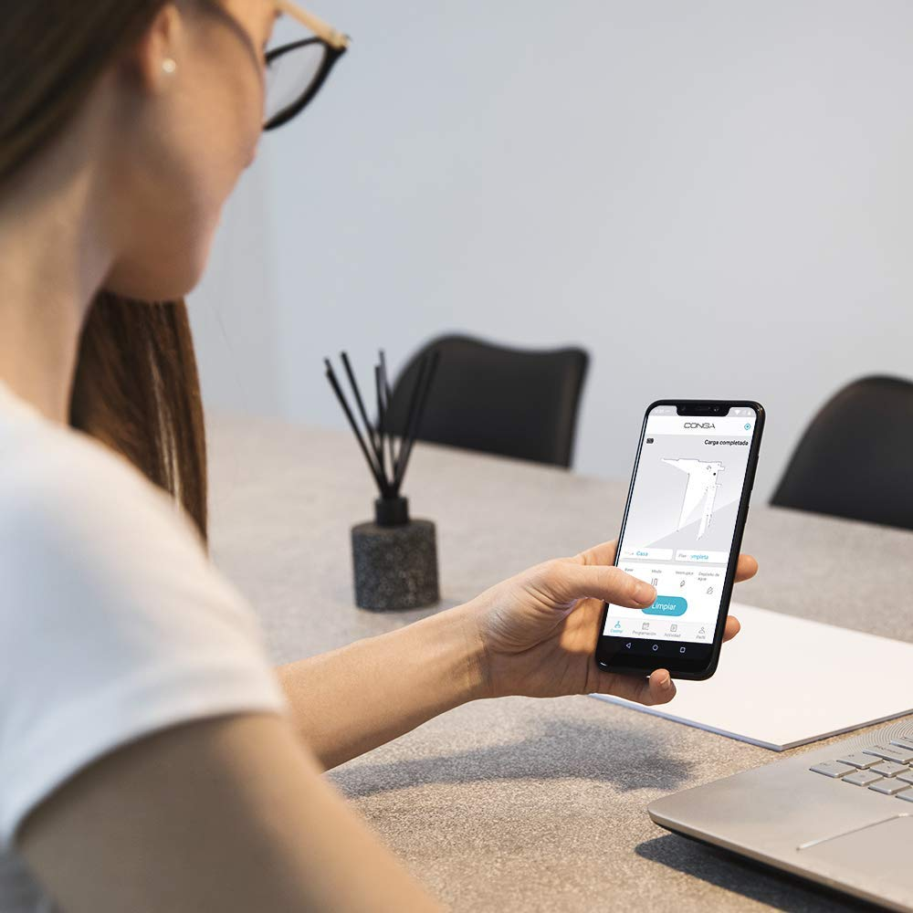 Una mujer sosteniendo un teléfono mostrando una app de limpieza. Escritorio con laptop y difusor de aroma. Fondo: oficina.