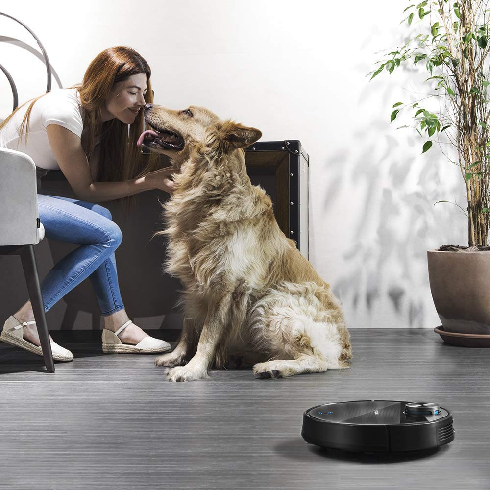 Una mujer y un perro golden retriever en una sala de estar. Una aspiradora robot negra en el suelo.