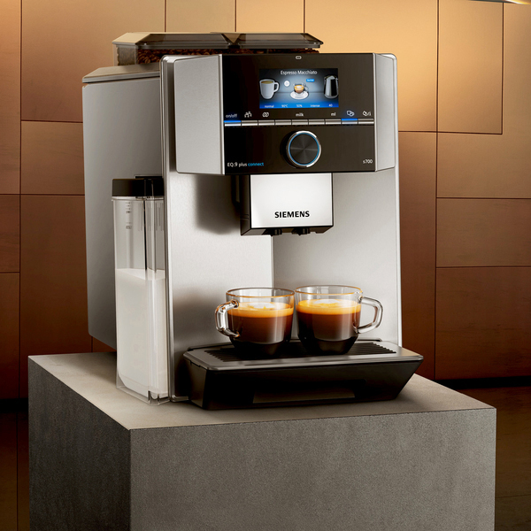 Silberne und schwarze Espressomaschine mit Touchscreen, zwei Kaffeetassen und einem Milchbehälter auf grauer Oberfläche.