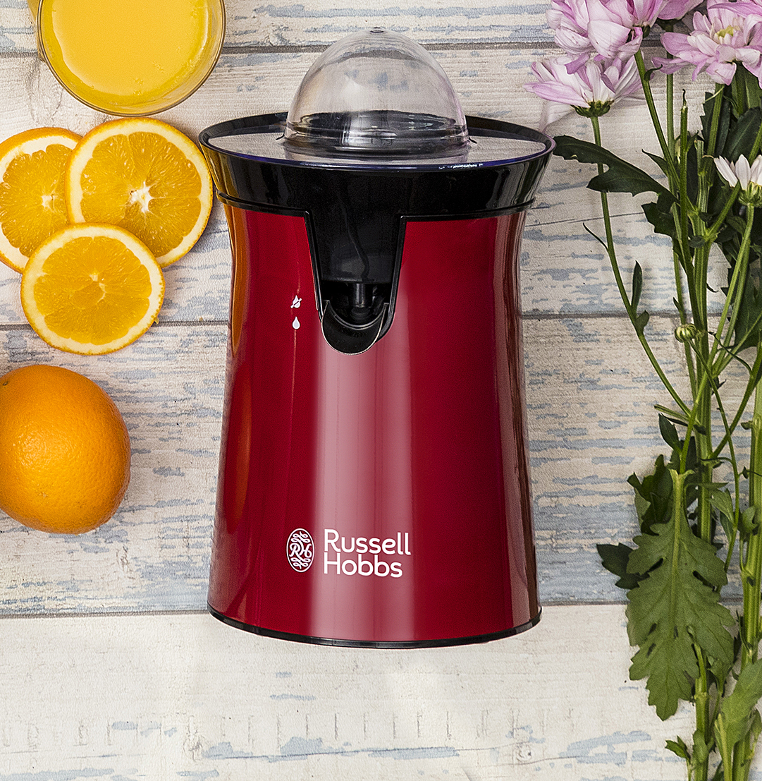 Piros Russell Hobbs gyümölcsprés narancsokkal, lével és rózsaszín virágokkal fa mintás felületen.