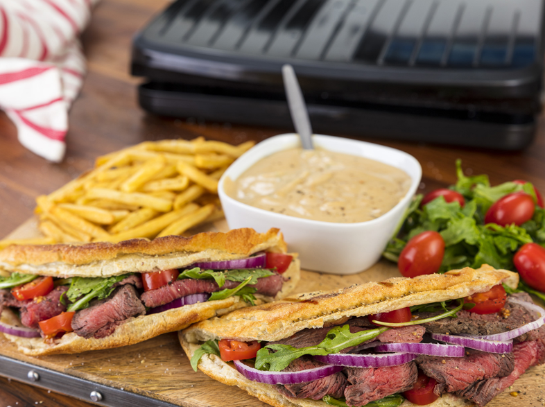 Két steak szendvics, sült krumpli, szósz és saláta egy fa deszkán. Grill és piros csíkos kendő a háttérben.