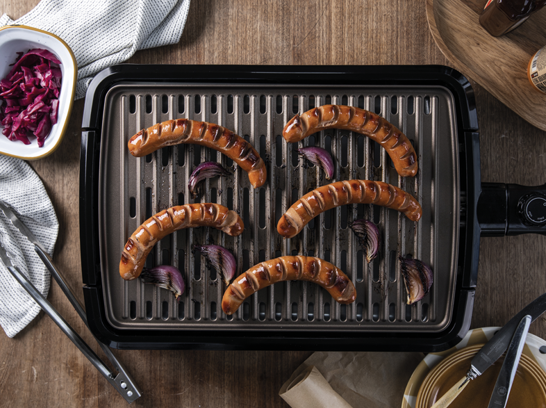 Kolbászok és vöröshagymák grillezése egy fekete grillen. Eszközök és köretek láthatók.