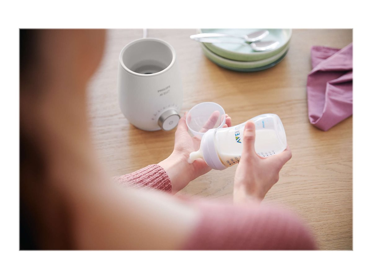Person gießt Milch in eine Babyflasche. Weißer Wärmer im Hintergrund.