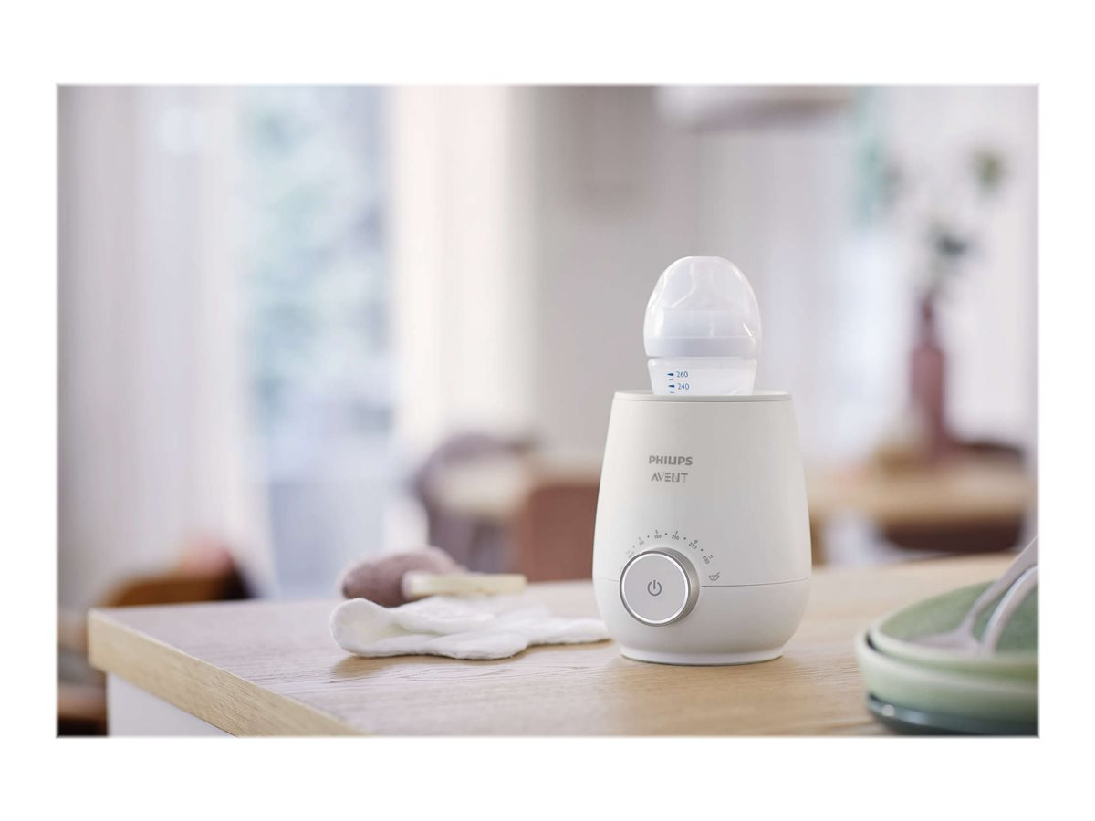 Weißer Babyflaschenwärmer. Flasche darin. Tisch mit Gegenständen. 'Philips Avent' sichtbar.