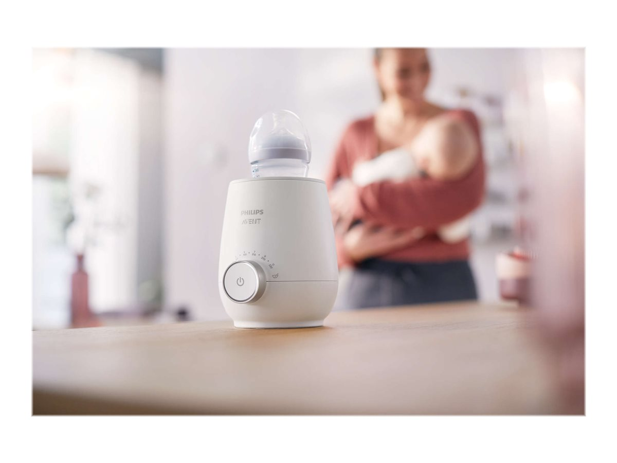 Ein weißer Babyflaschenwärmer mit einer Flasche obenauf und eine Frau, die im Hintergrund ein Baby hält.
