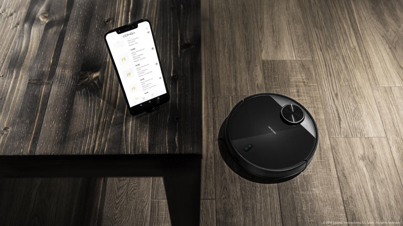 Un smartphone y un aspirador robótico negro en un suelo de madera. El teléfono tiene una aplicación abierta.