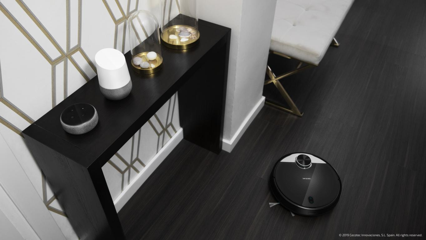 Una mesa negra con dispositivos domésticos inteligentes y un robot aspirador en el suelo.