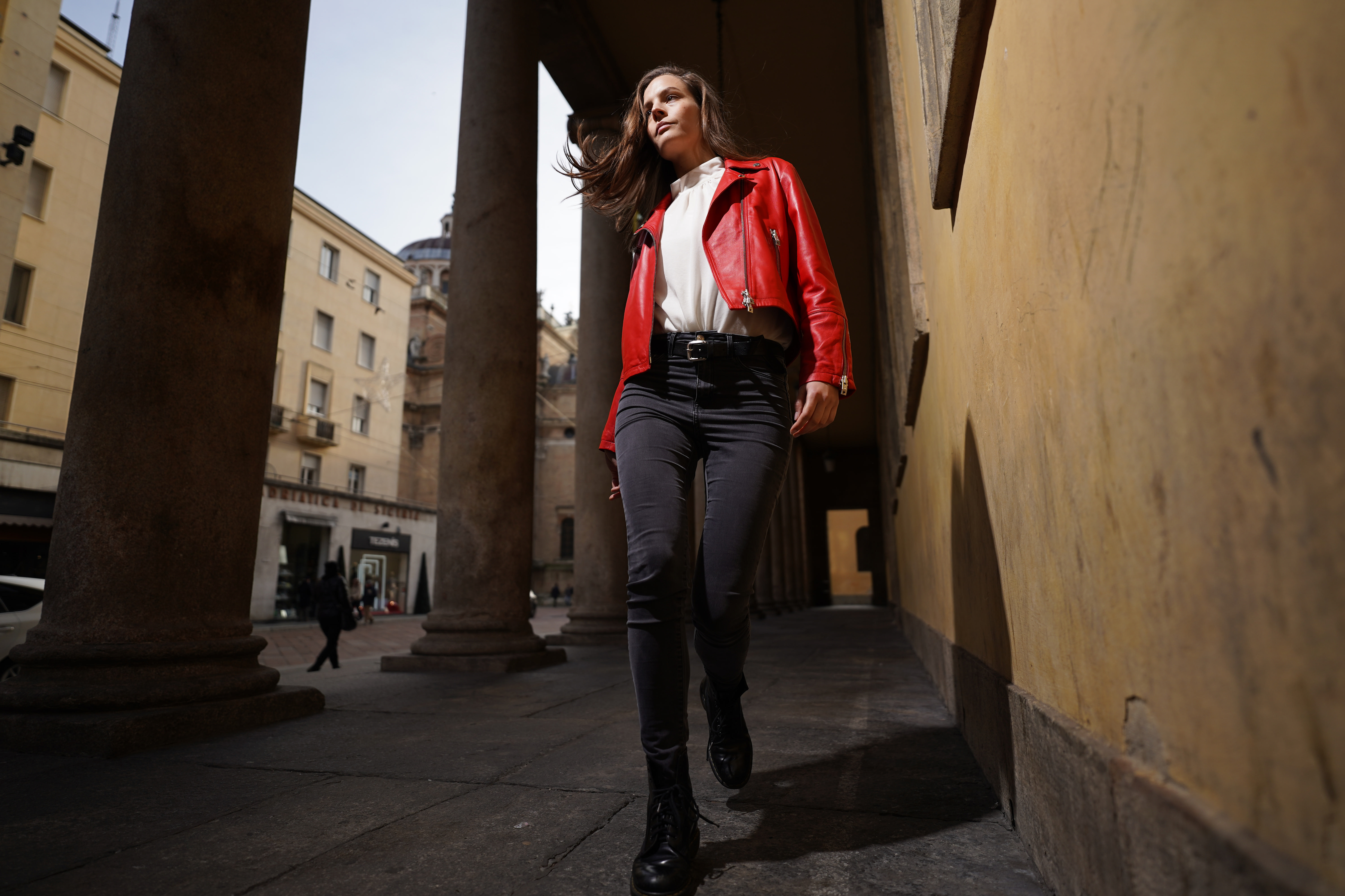 Eine Frau in roter Jacke geht auf die Kamera zu. Gebäude mit Säulen im Hintergrund.