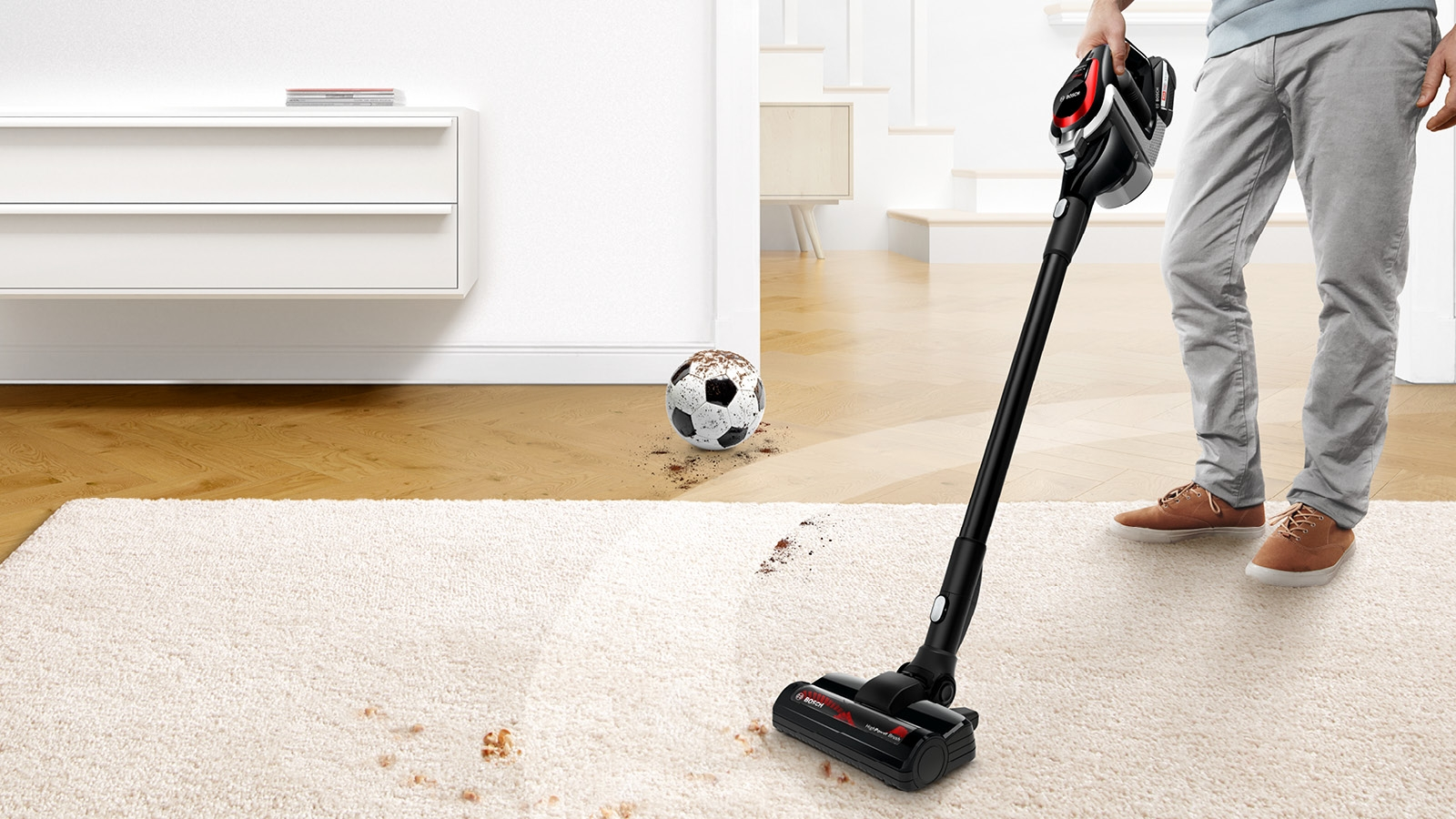 Person saugt Teppich mit einem kabellosen Staubsauger. Schmutz und Fußball sichtbar.