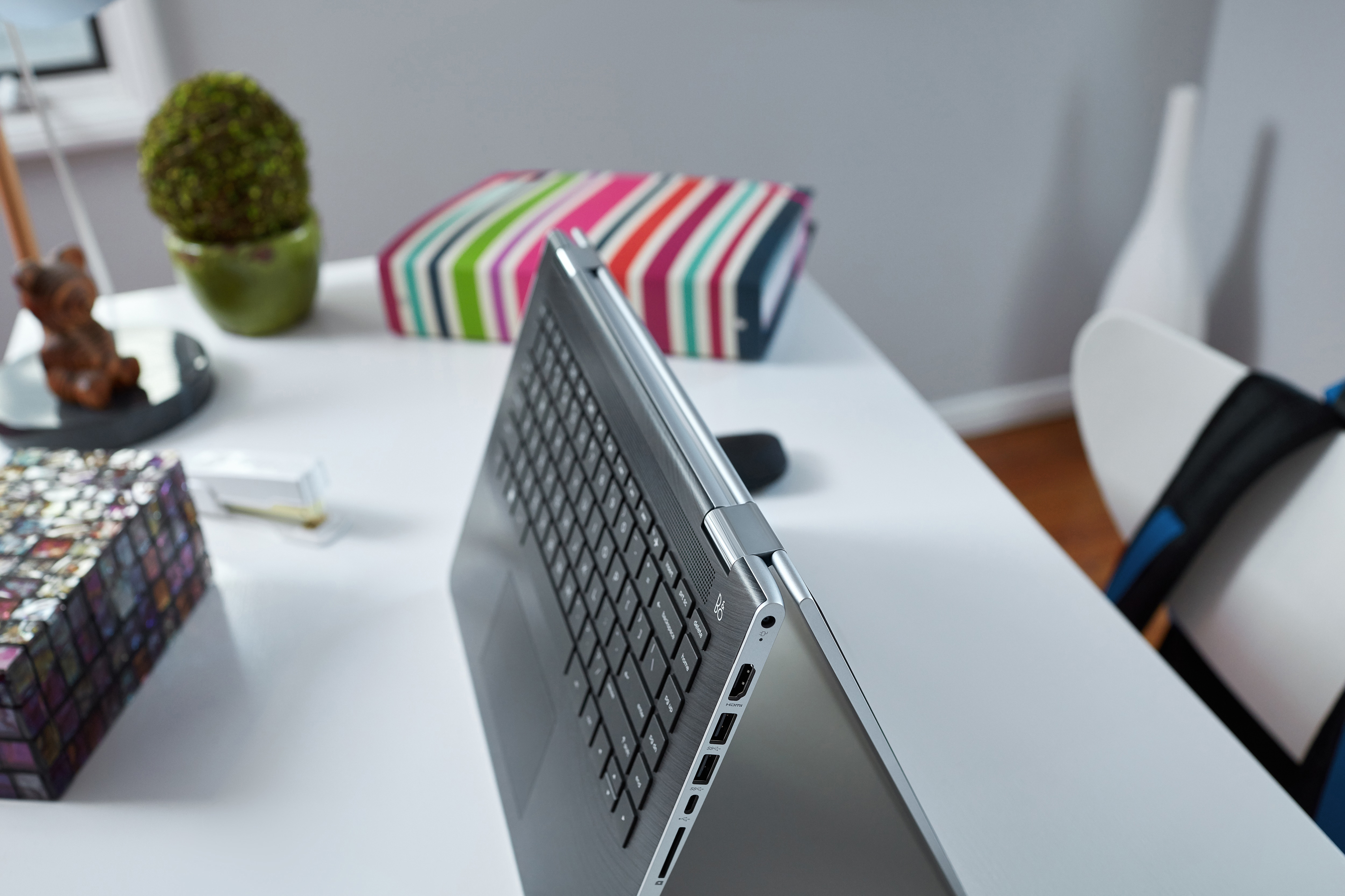 Nahaufnahme eines silbernen Laptops im Zeltmodus auf einem weißen Schreibtisch, mit einer bunten Schachtel und einer Pflanze.