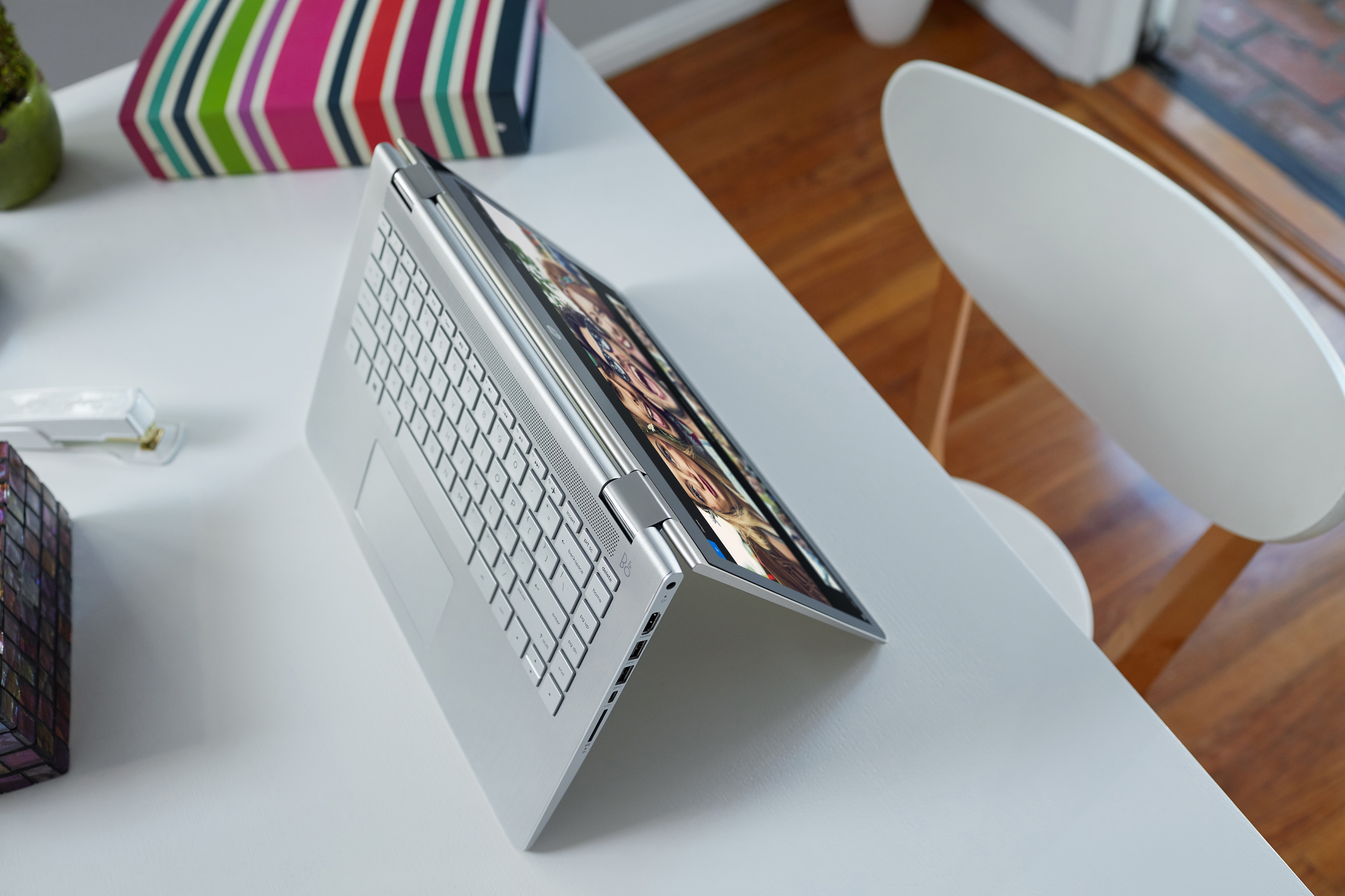 Ein silberner Laptop im Zeltmodus steht auf einem weißen Tisch neben einem bunten Hefter und einem weißen Stuhl.