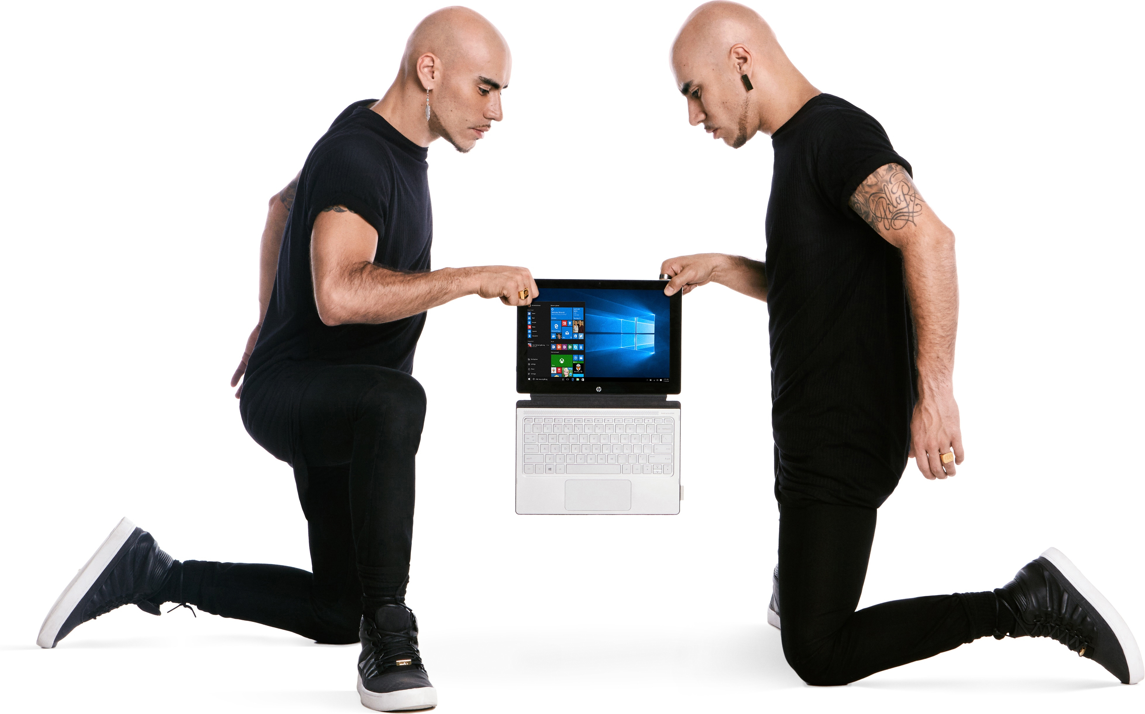 Zwei identische kahlköpfige Männer in schwarzer Kleidung knien und halten einen Laptop vor weißem Hintergrund.
