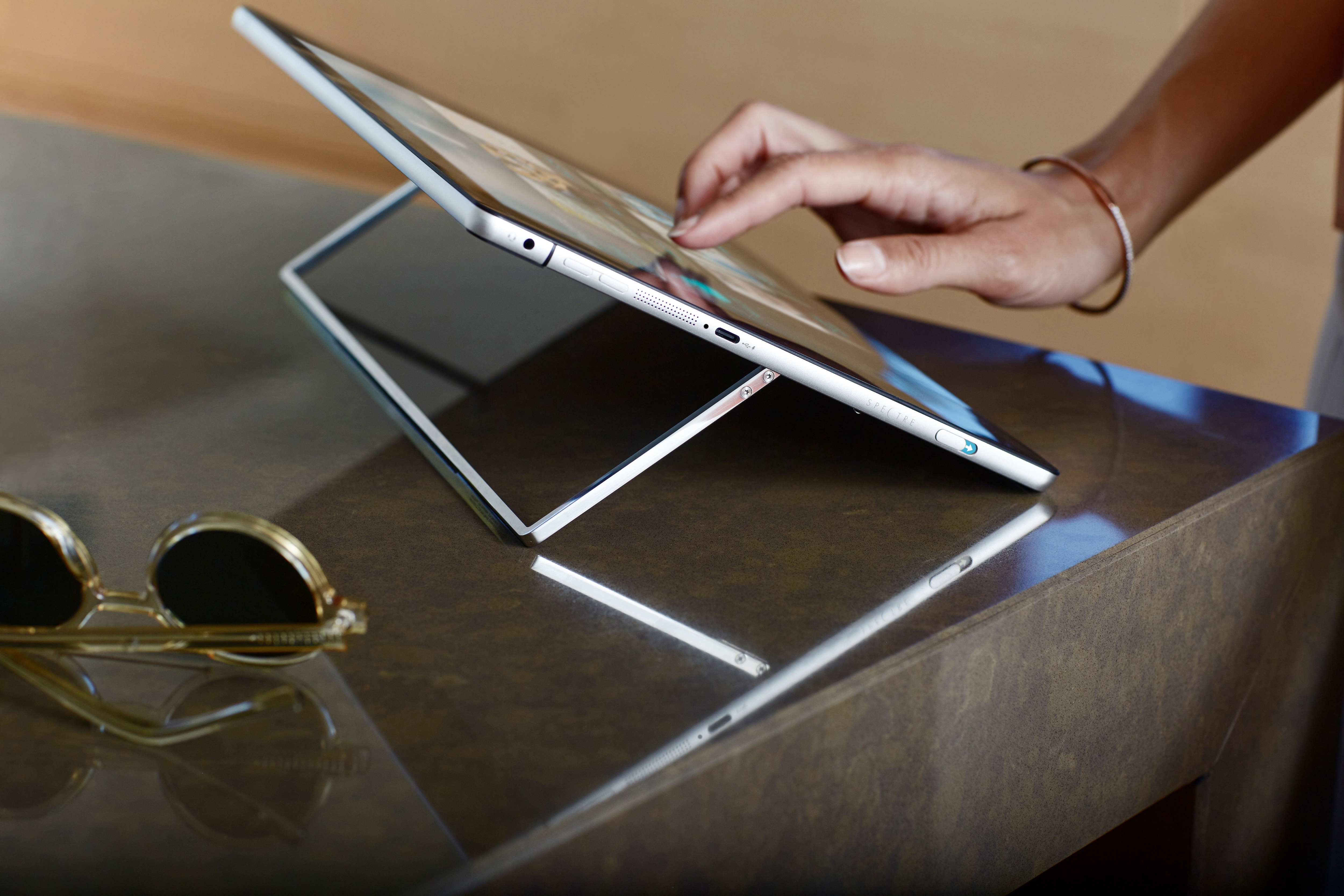 Eine Hand tippt auf einem Tablet im Ständer auf einem glänzenden Tisch, Sonnenbrille links.