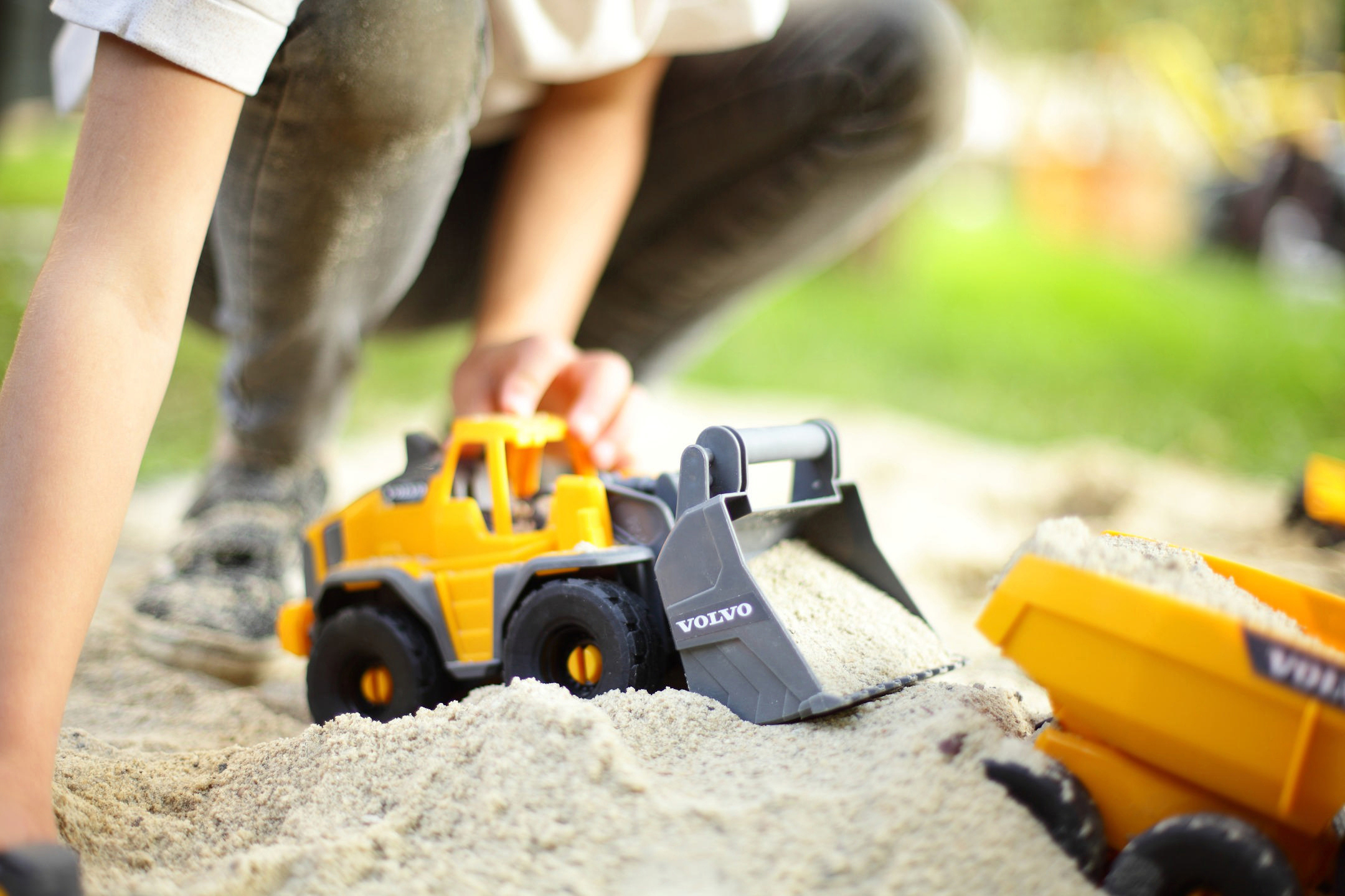 Ein Kind spielt mit einem gelben Volvo-Spielzeugbagger im Sand, ein weiterer Spielzeug-LKW im Hintergrund.