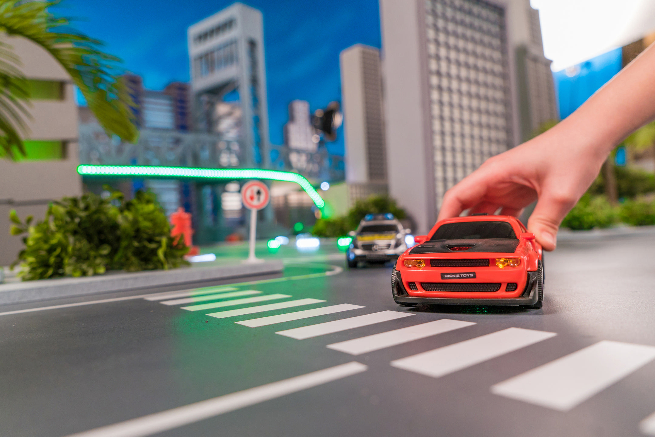 Eine Hand platziert ein rotes Spielzeugauto auf einem Zebrastreifen. Ein Polizeiauto steht dahinter. Stadtmodelle und eine Palme sind im Hintergrund.