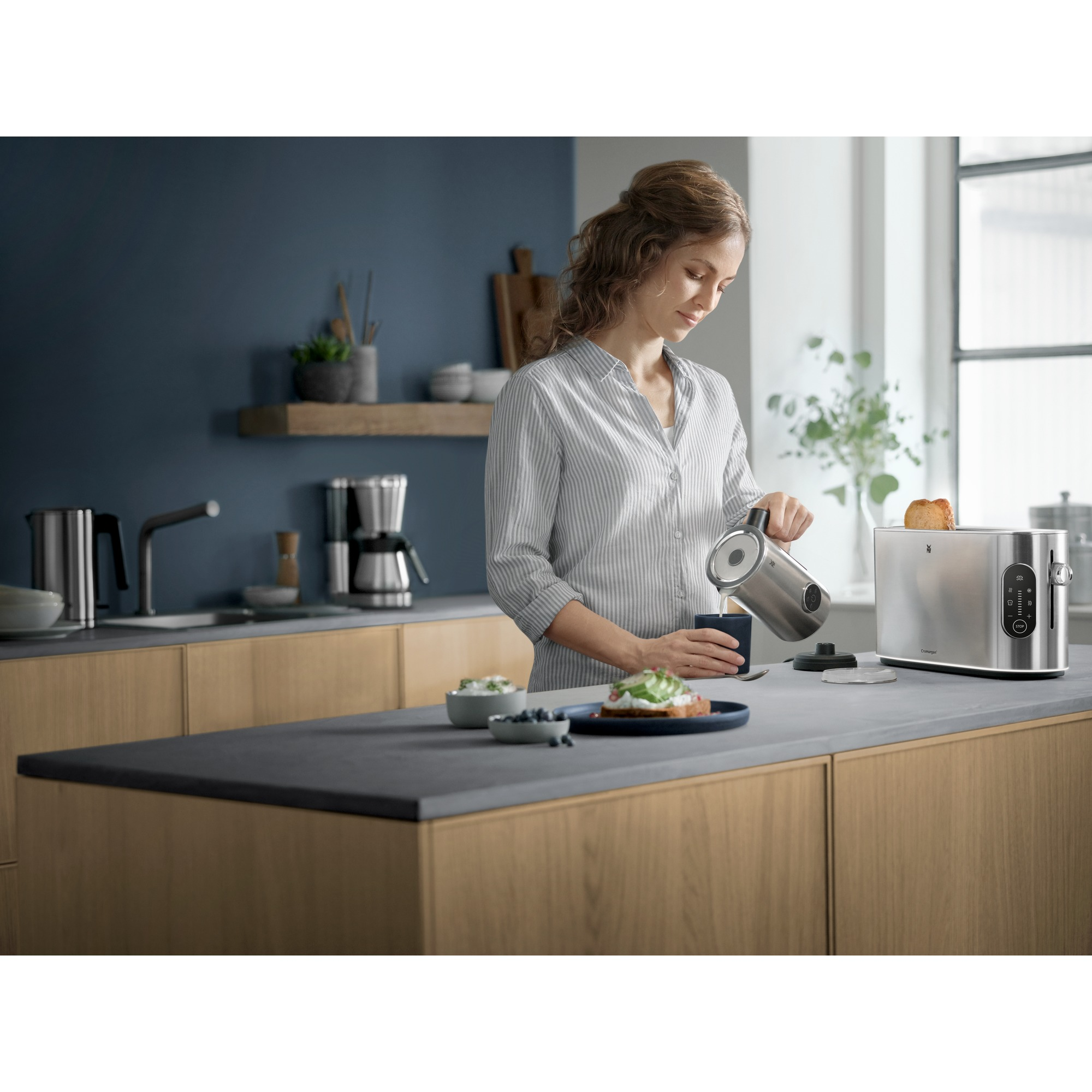 Woman pouring water into a cup near a toaster and food on a counter.