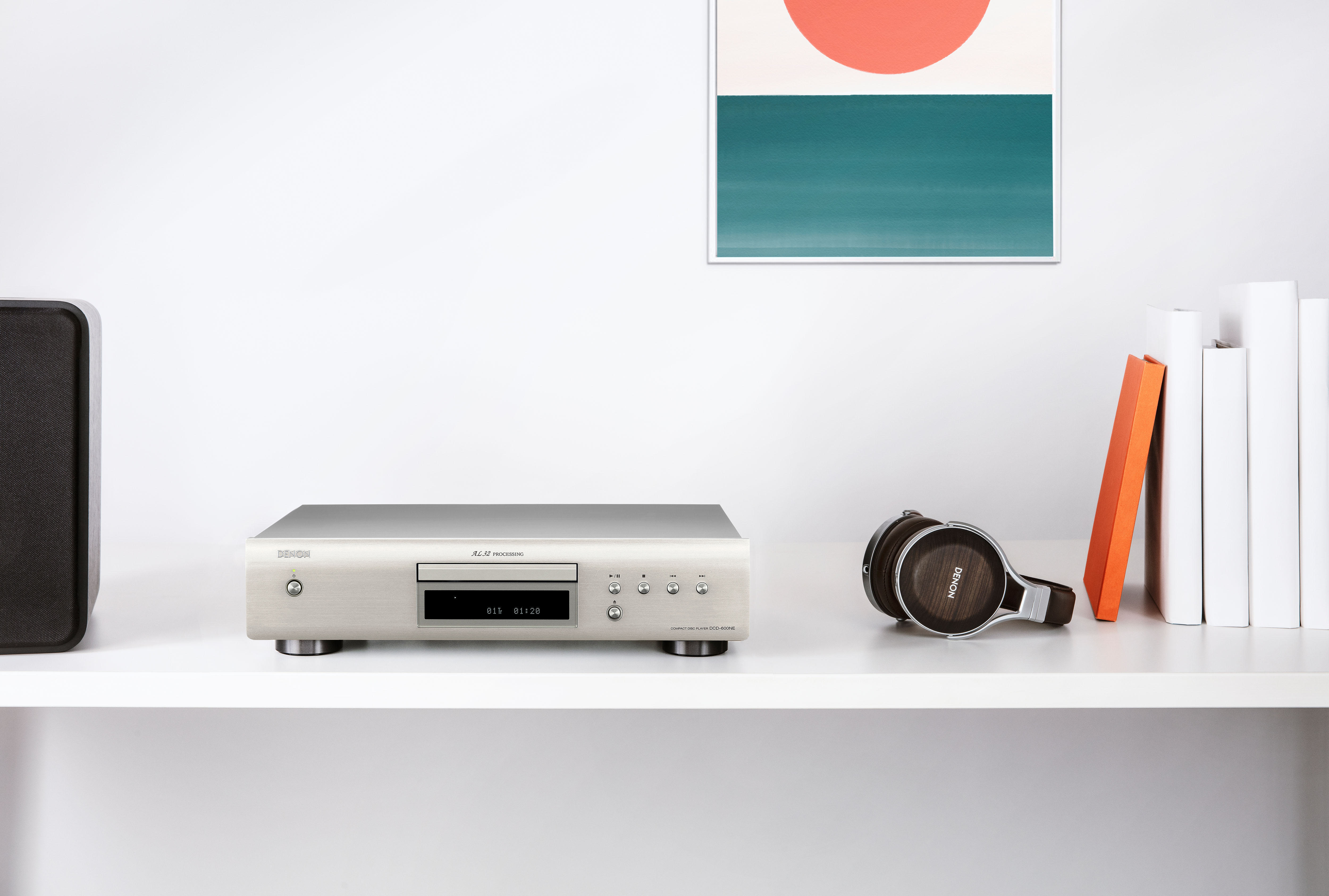 Ein silberner Denon CD-Player auf einem weißen Regal mit Kopfhörern, Büchern und Kunst im Hintergrund.