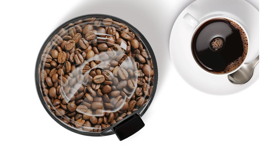 Vista cenital de un molinillo de café con granos de café y una taza de café sobre fondo blanco.