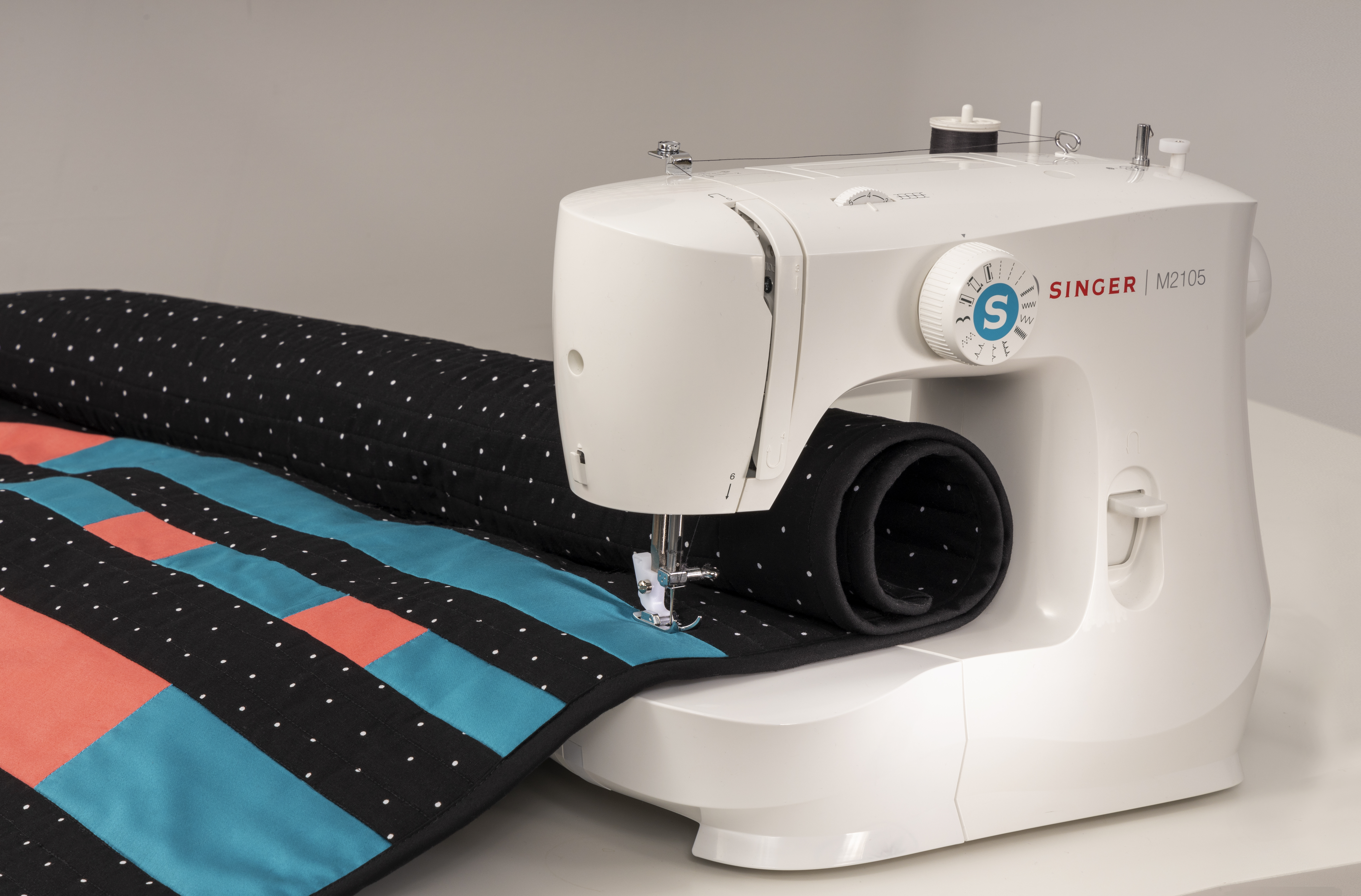 Sewing machine sewing a quilt. The quilt is a patchwork of black, coral and teal colors.