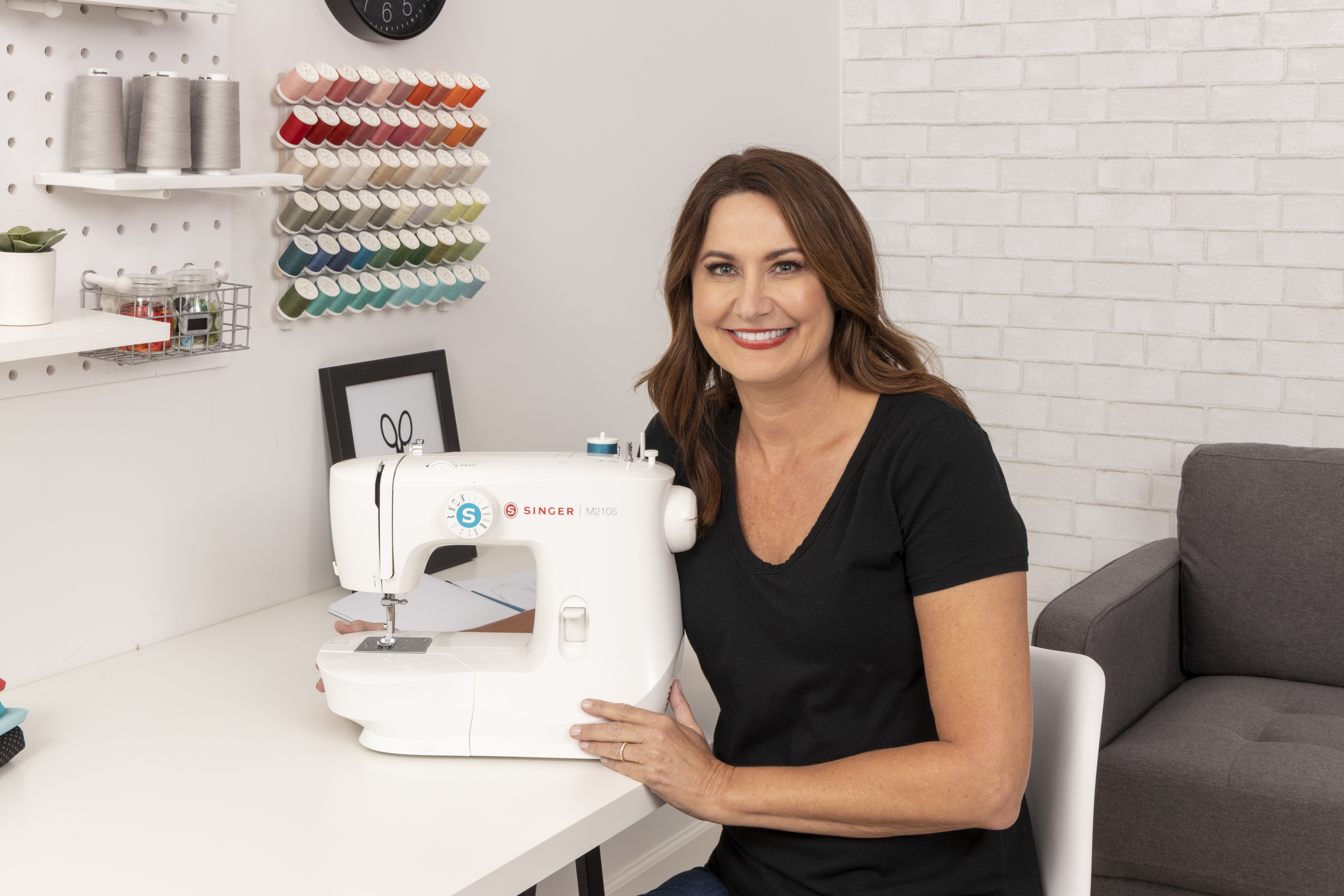 A woman smiling next to a white sewing machine. She is in a well-lit room with spools of thread.