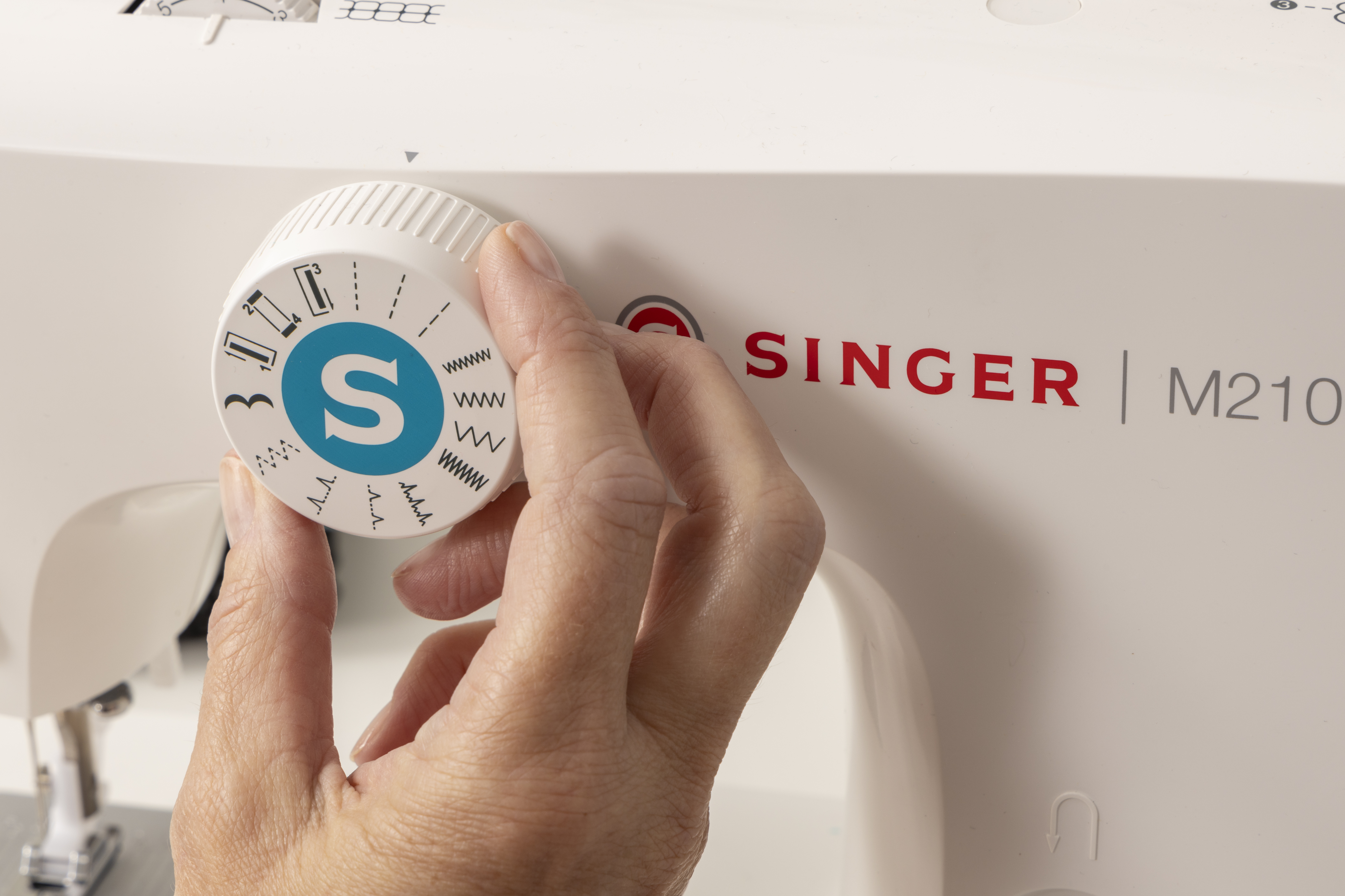 A hand adjusting the stitch selector dial on a white Singer M210 sewing machine.