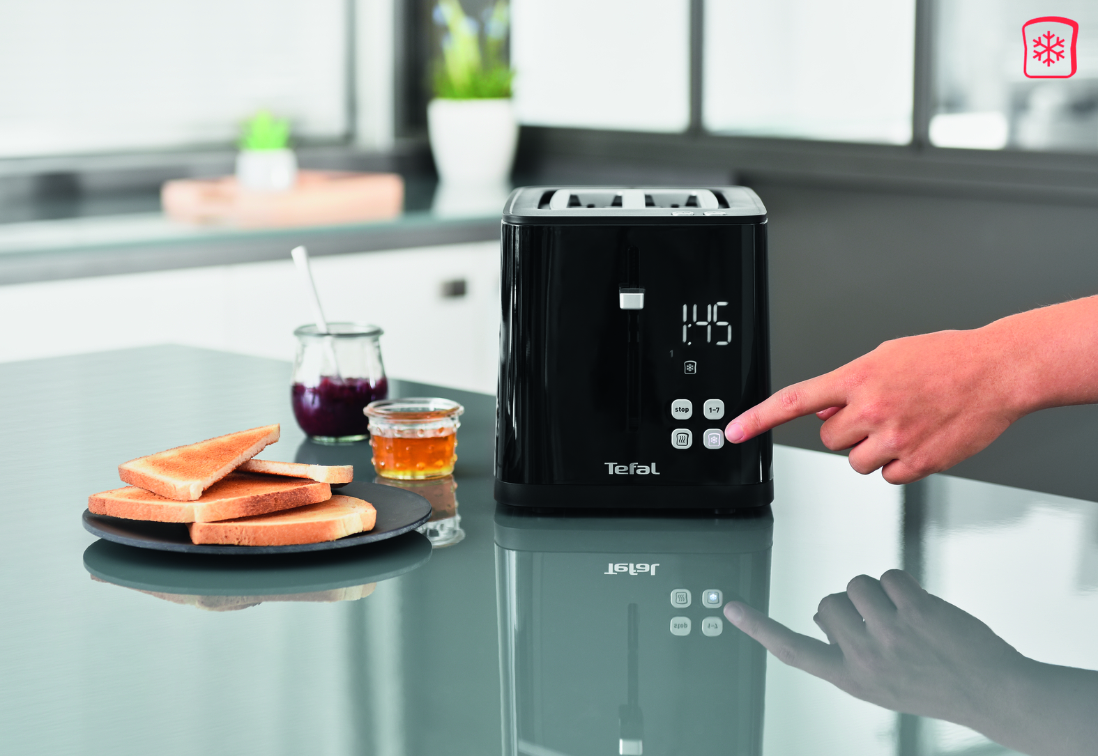 Een zwarte Tefal broodrooster met een hand die op een knop drukt. Toast en jam staan op het aanrecht.