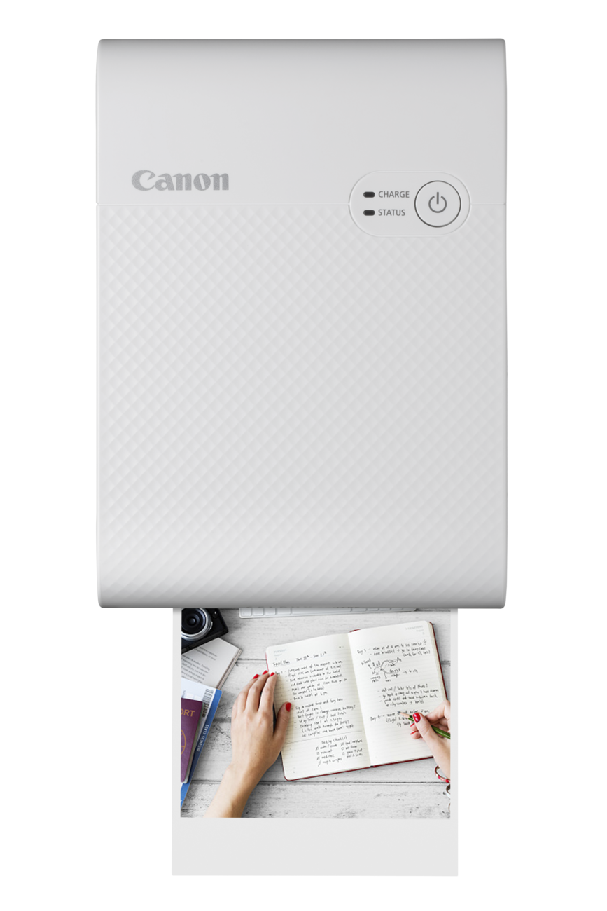 A white Canon Selphy photo printer with a person writing in a notebook, and a photo being printed.
