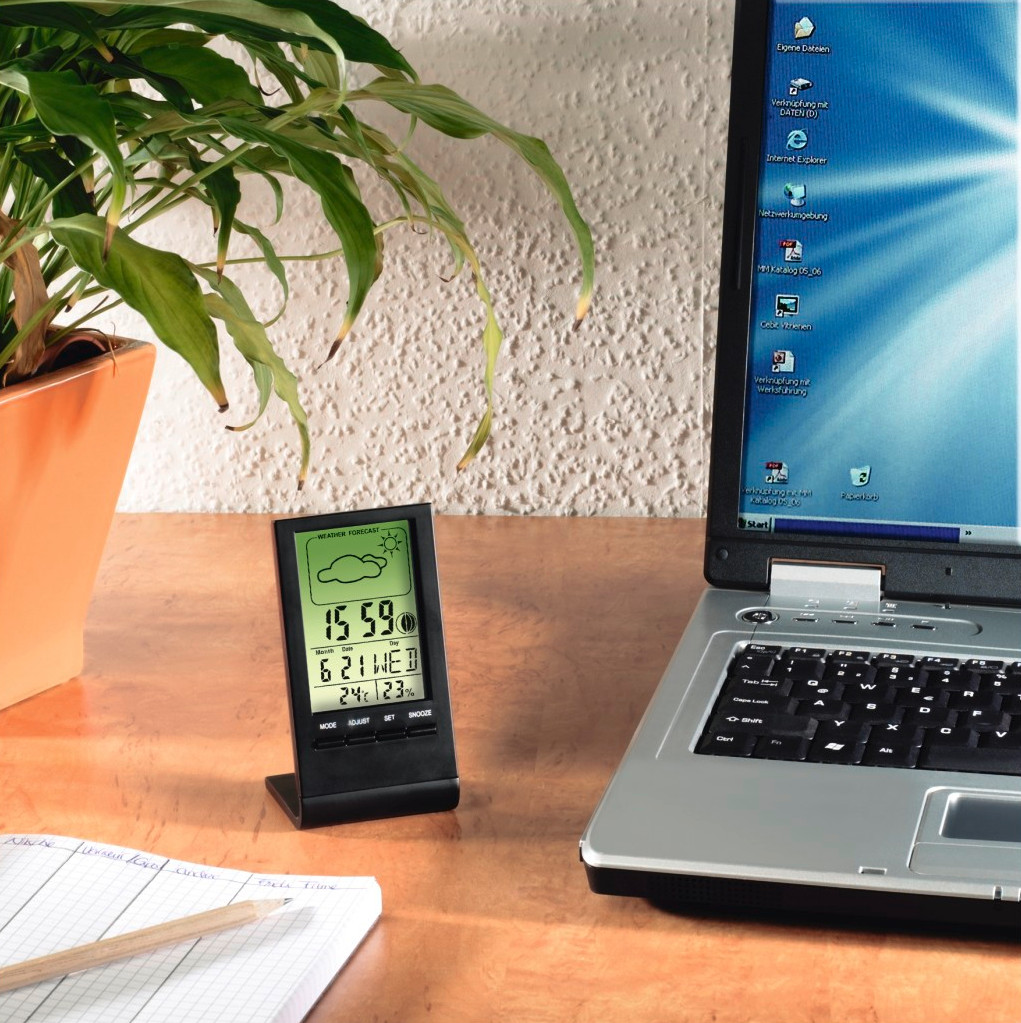 Digital weather station on a desk next to a laptop and a potted plant. The screen shows time and weather forecast.