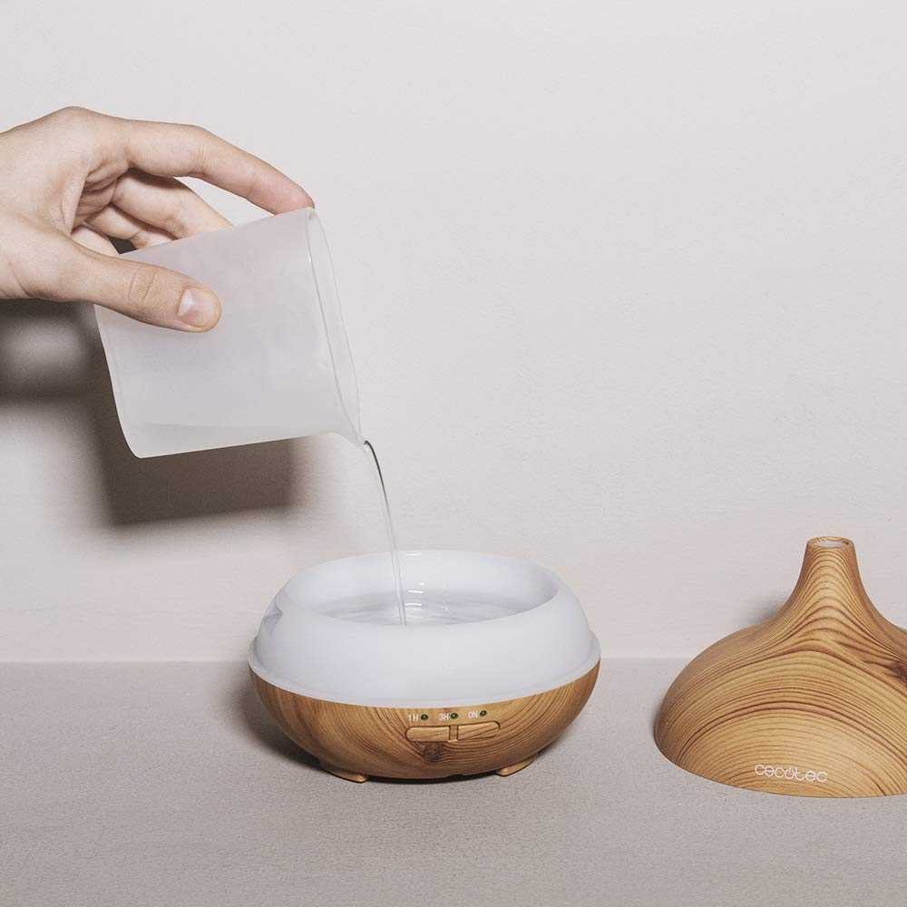 Mano vertiendo agua de un recipiente transparente en un difusor con aspecto de madera. La tapa está al lado.