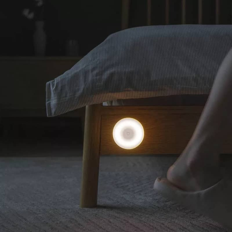 Una lámpara de noche redonda y brillante está debajo de una cama. Se ve un pie con una zapatilla.