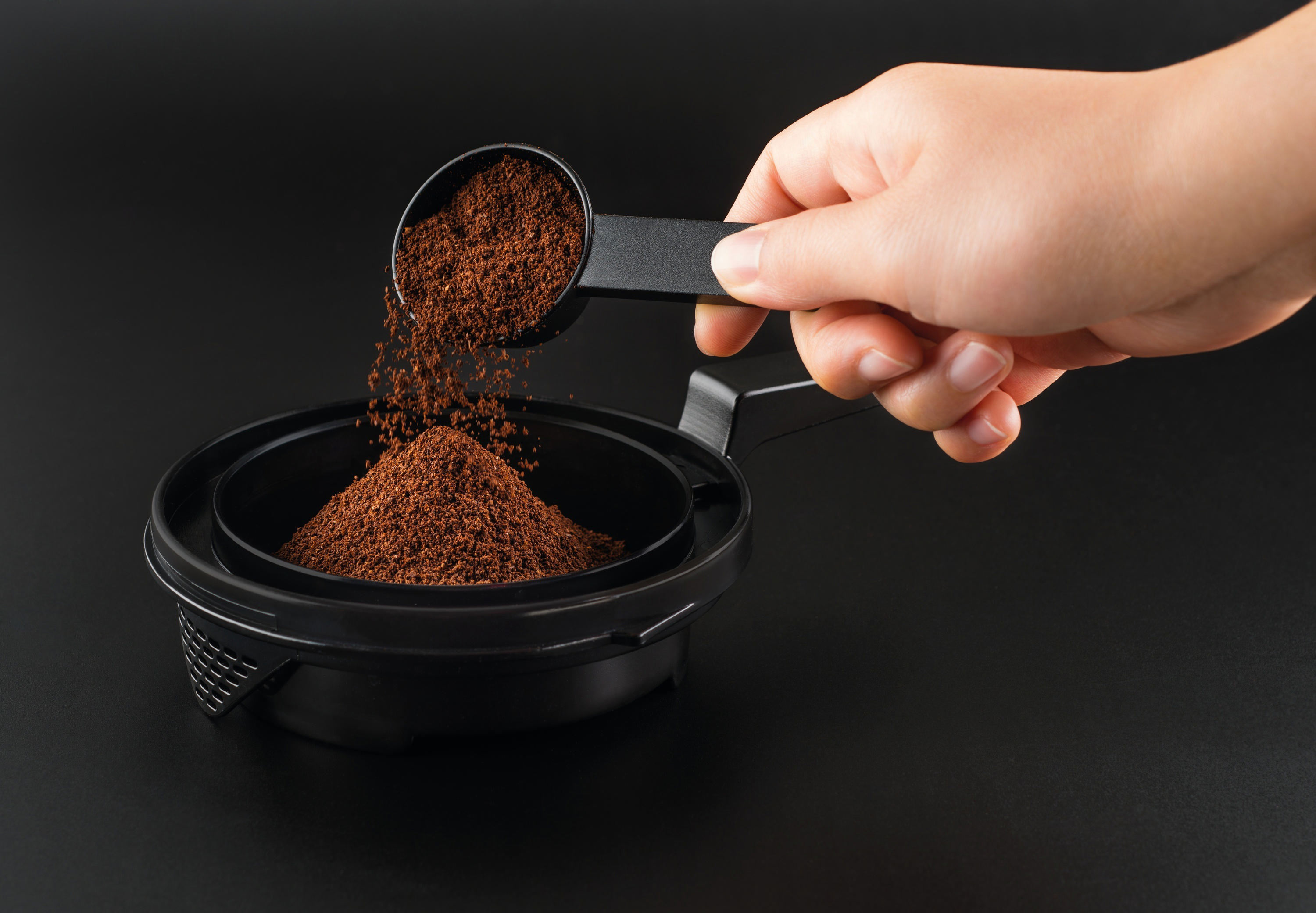 Hand gießt Kaffeepulver aus einem Löffel in einen schwarzen Filter.