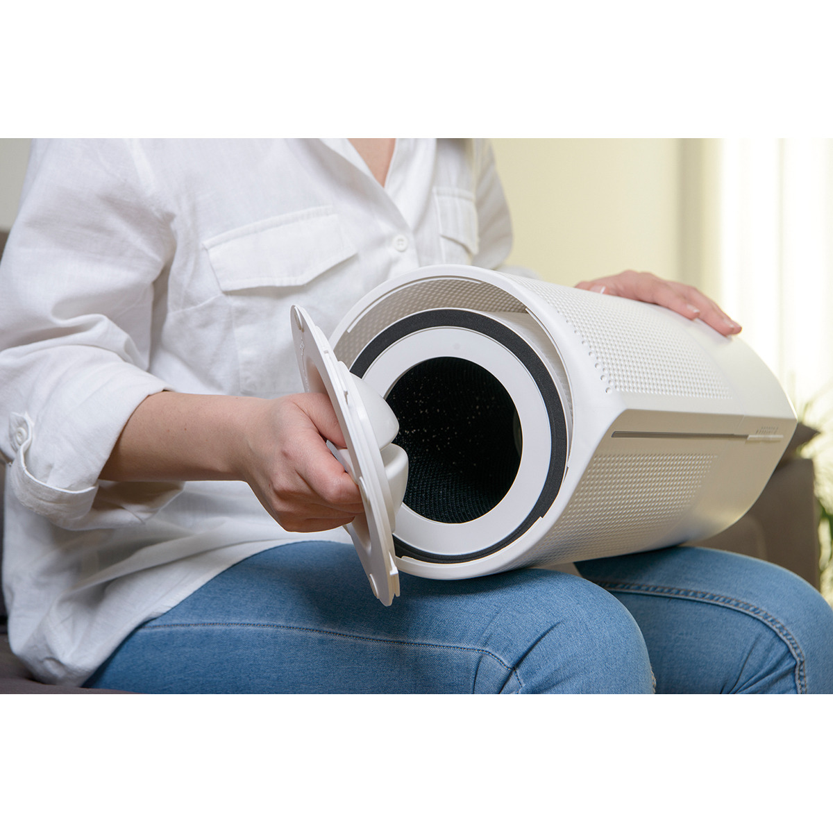 Persona abriendo compartimento de filtro de purificador de aire blanco, sentada en un sofá.