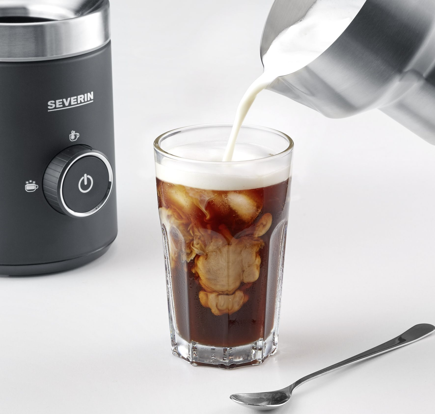 Milch wird in ein Glas Eiskaffee gegossen. Eine schwarze Kaffeemaschine und ein Löffel sind ebenfalls vorhanden.