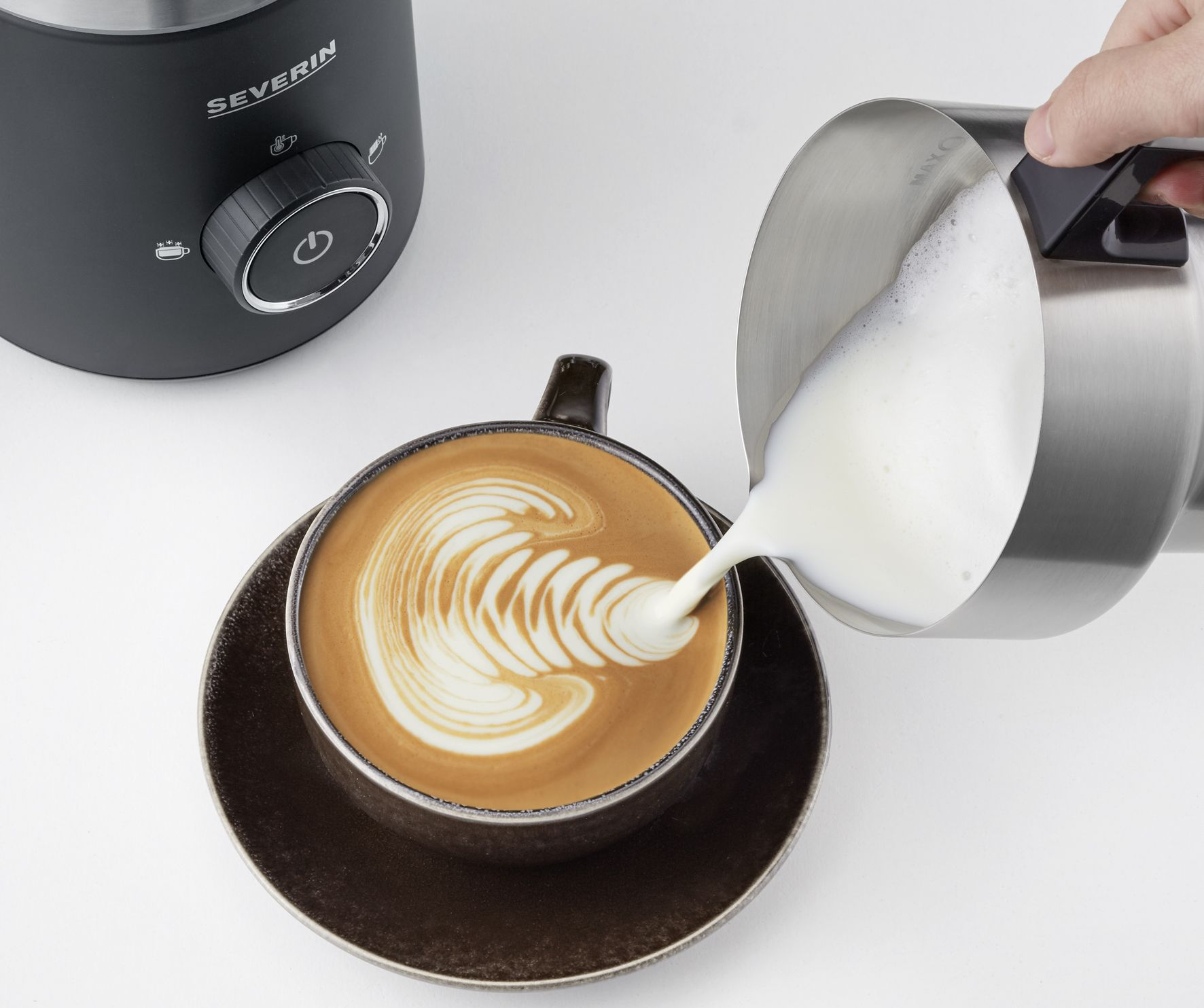 Eine Hand gießt Milch aus einem Metallkrug in eine Kaffeetasse mit Latte Art, in der Nähe eines schwarzen Milchaufschäumers, auf weißem Untergrund.