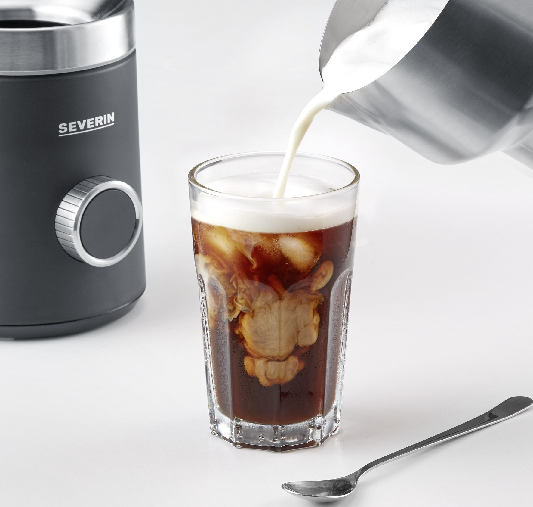 Milch wird in ein Glas Eiskaffee gegossen. Eine Kaffeemühle, ein Löffel und eine weiße Oberfläche vervollständigen die Komposition.