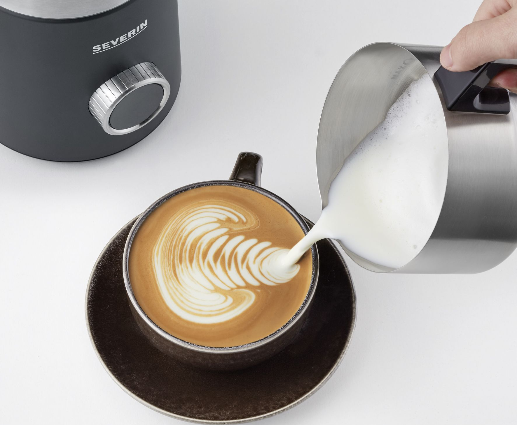 Eine Hand gießt geschäumte Milch in eine Kaffeetasse mit Latte Art. Im Hintergrund ein Milchaufschäumer.