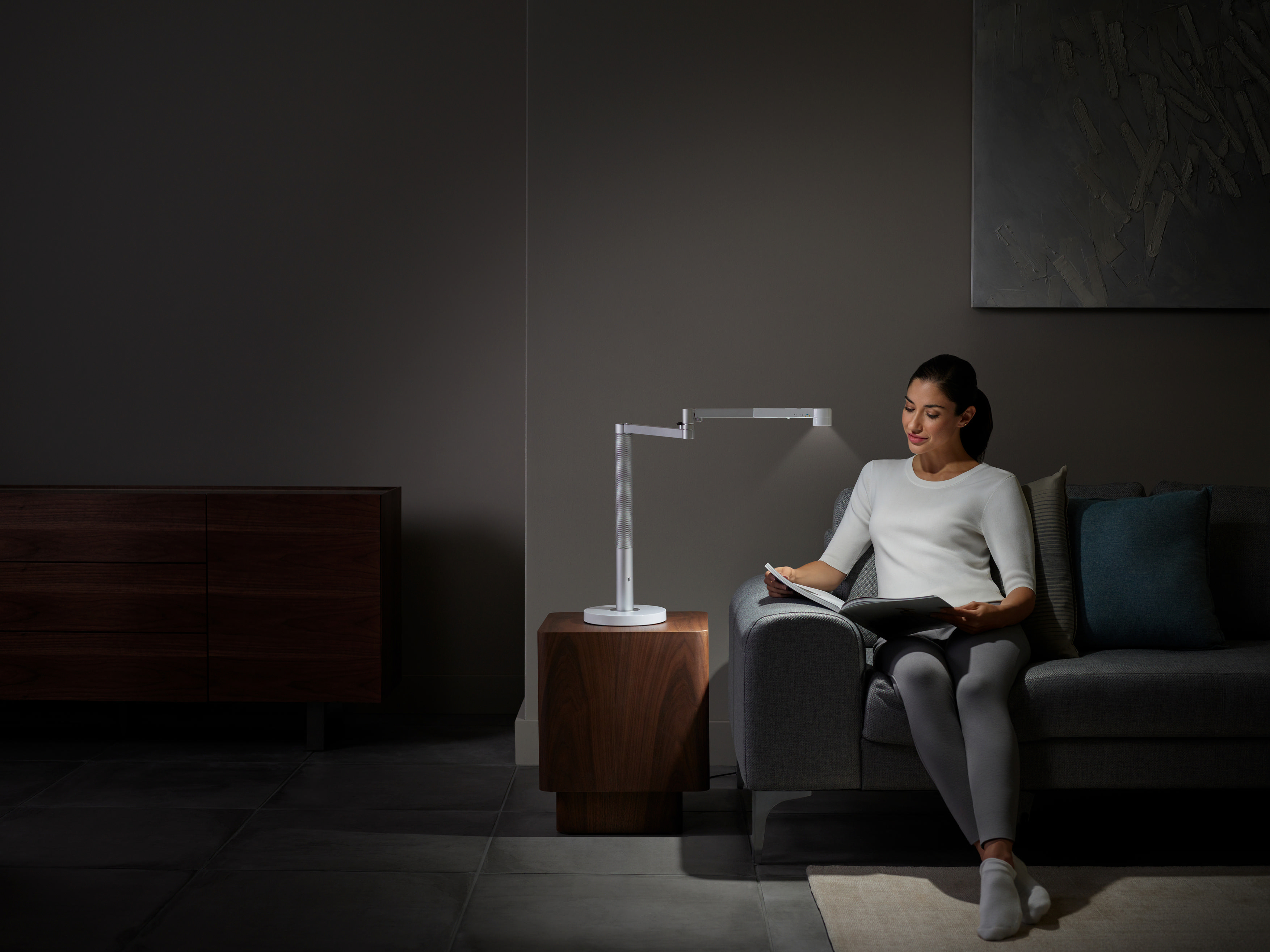 Eine Frau liest unter einer weißen Schreibtischlampe. Sie sitzt auf einem grauen Sofa neben einem hölzernen Beistelltisch.