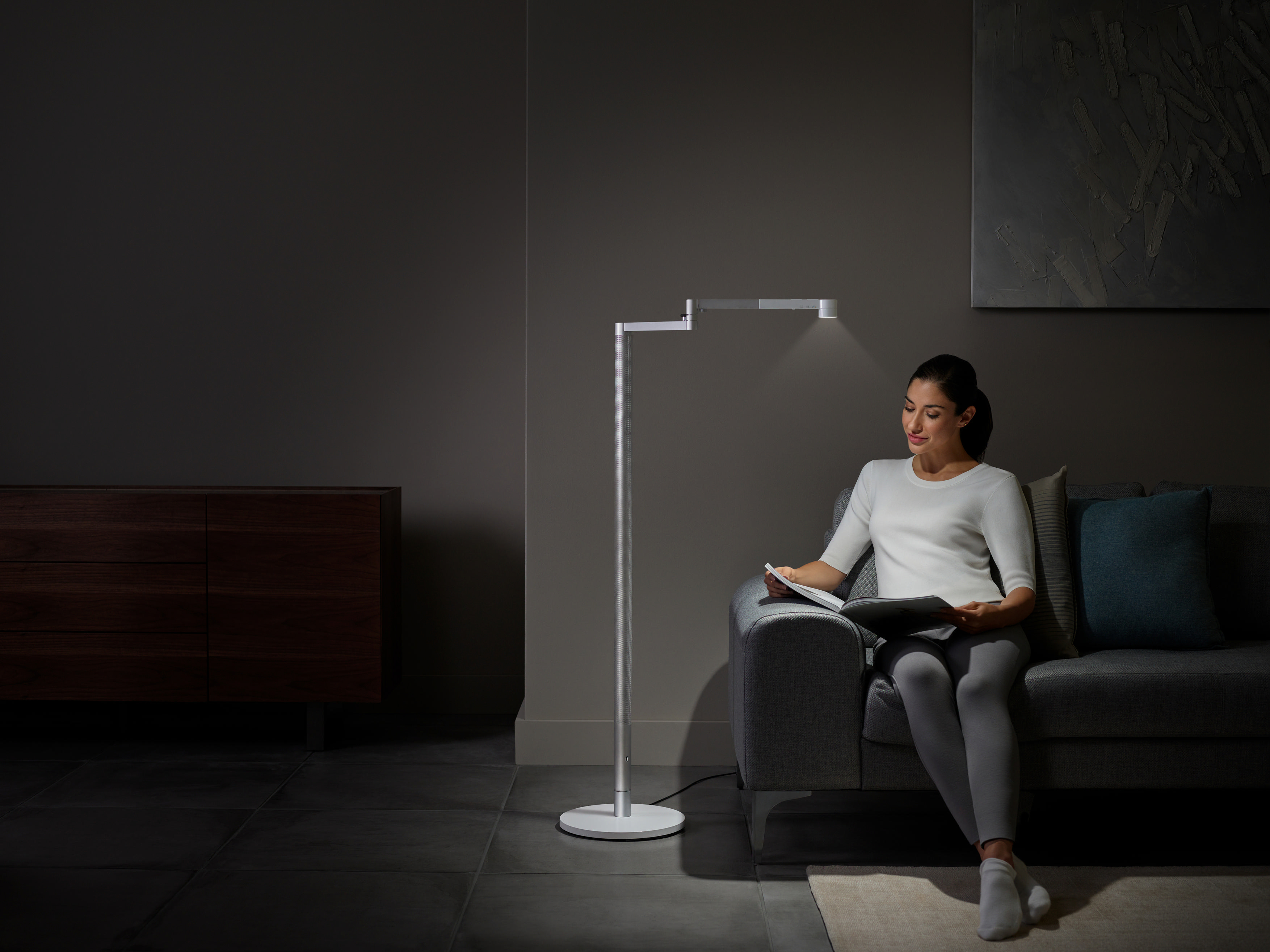 Frau liest unter einer modernen Stehlampe, sitzt auf einem grauen Sofa in dunklem Raum.